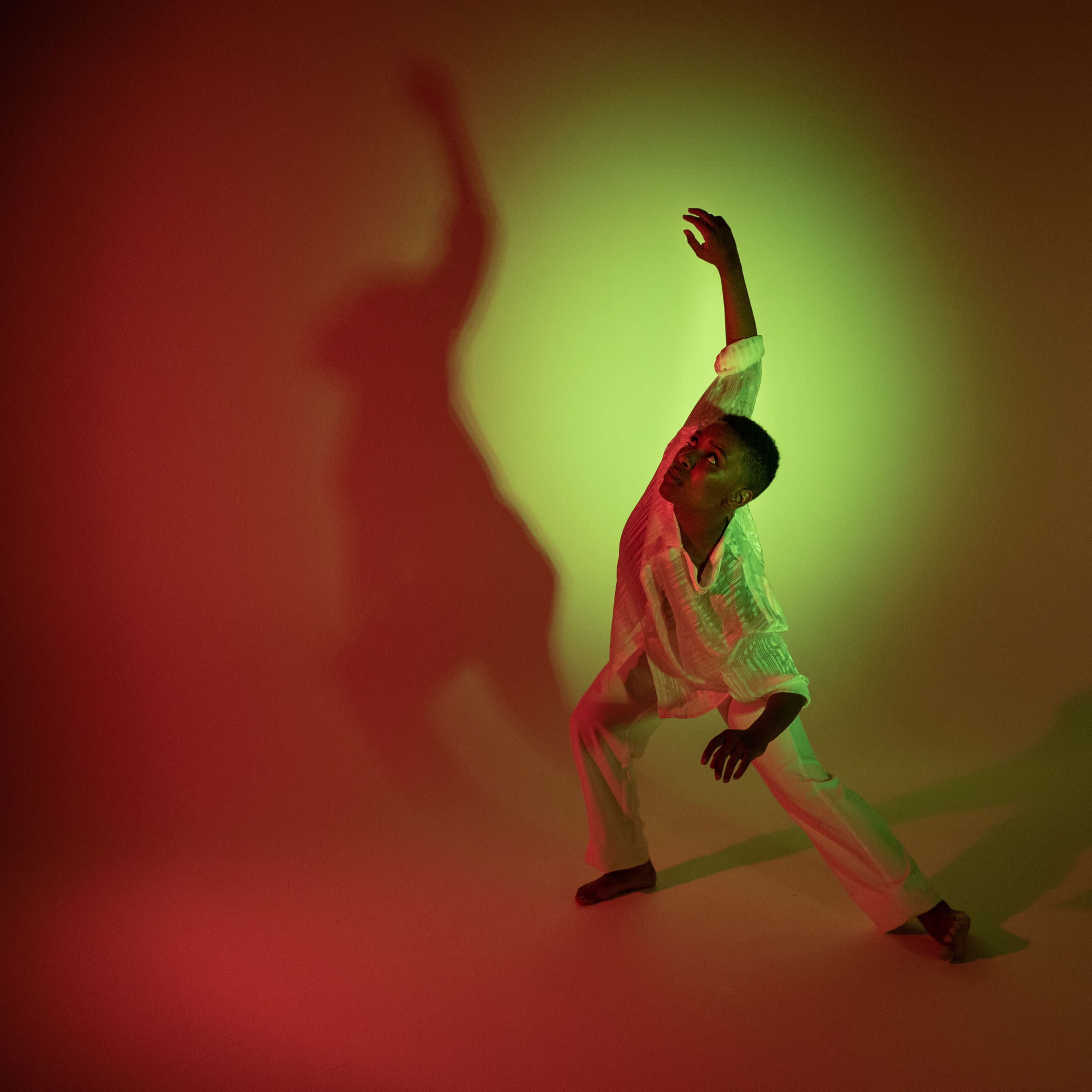 A dancer poses dramatically against a backdrop illuminated in red and green, casting elongated shadows on the surface.