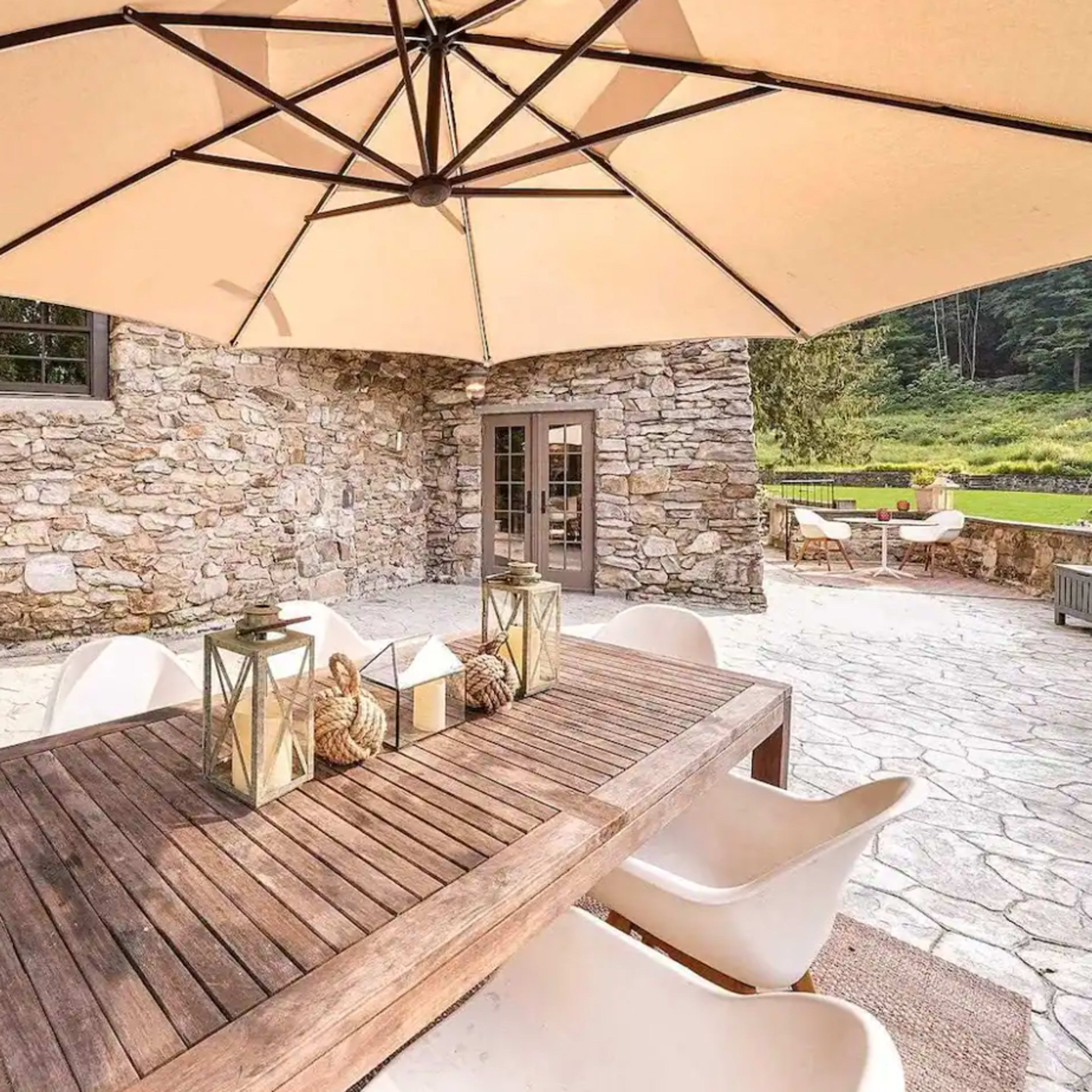 A wooden dining table with white chairs is set under a large umbrella on a stone patio beside a rustic stone wall.