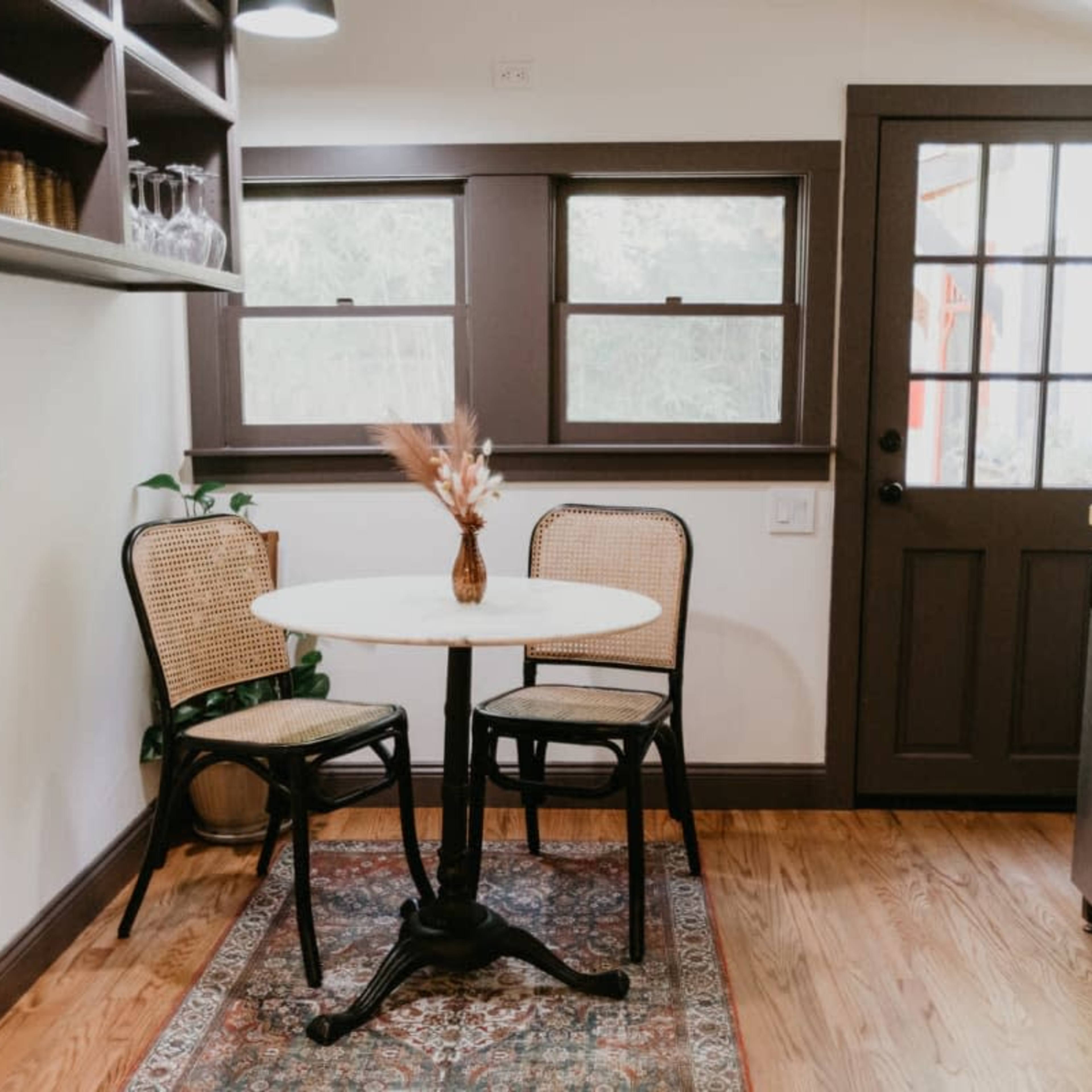 A small dining area features a round table with two chairs, a vase of dried flowers, a patterned rug, and a doorway leading to the outside.