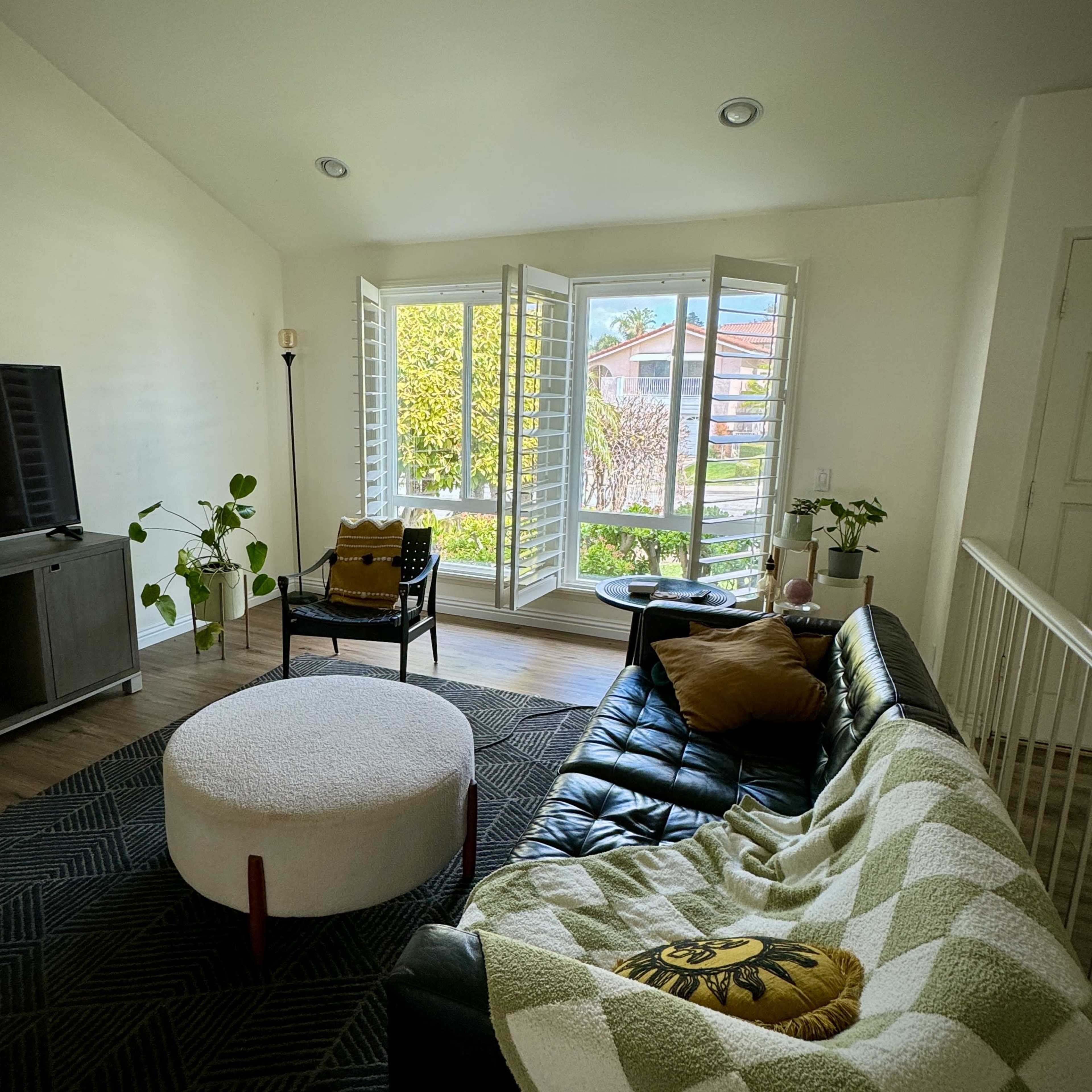 A bright living room with a large window, a black leather sofa, a round white coffee table, and various plants.