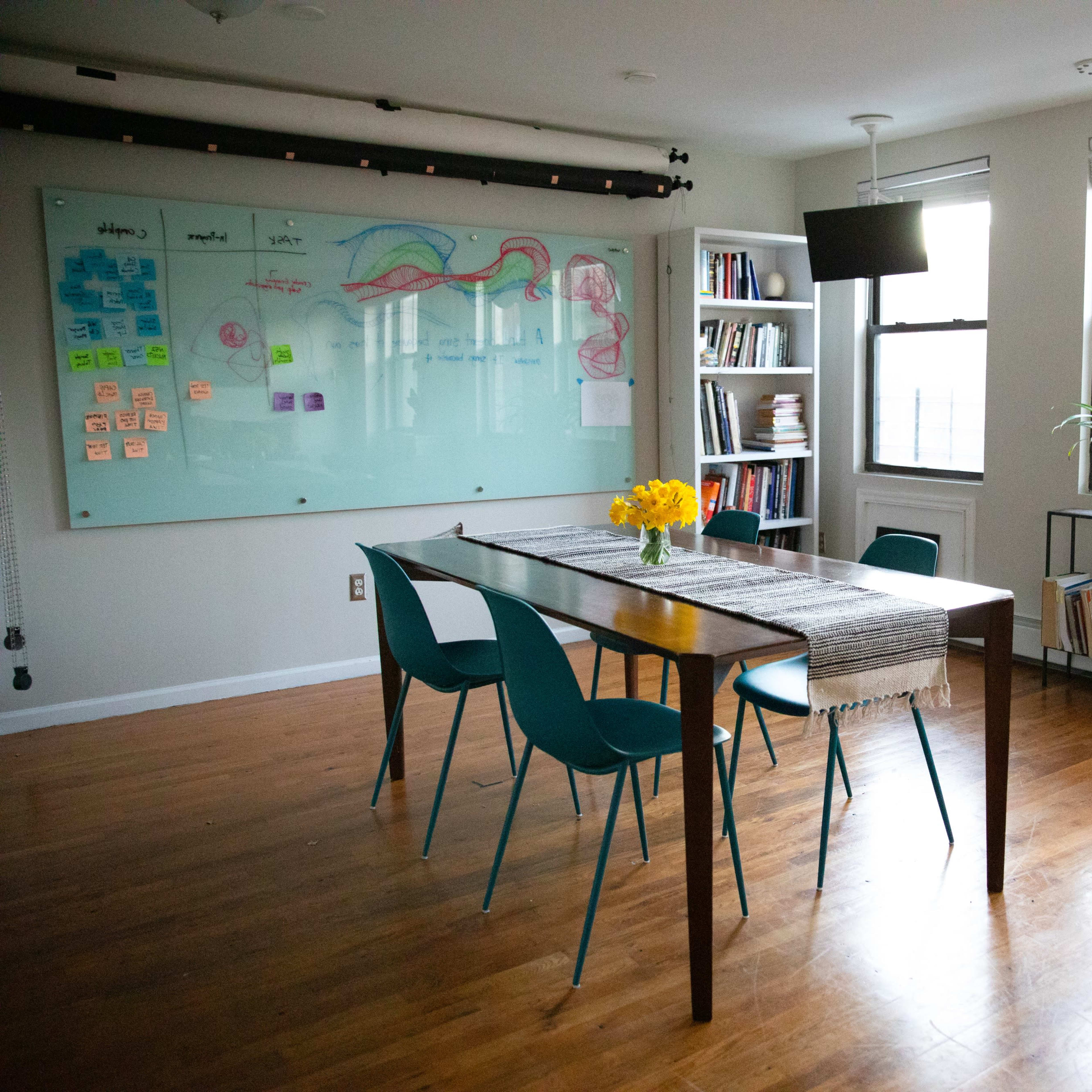 A bright, organized room with a wooden table surrounded by teal chairs, a whiteboard covered in notes, and shelves filled with books.