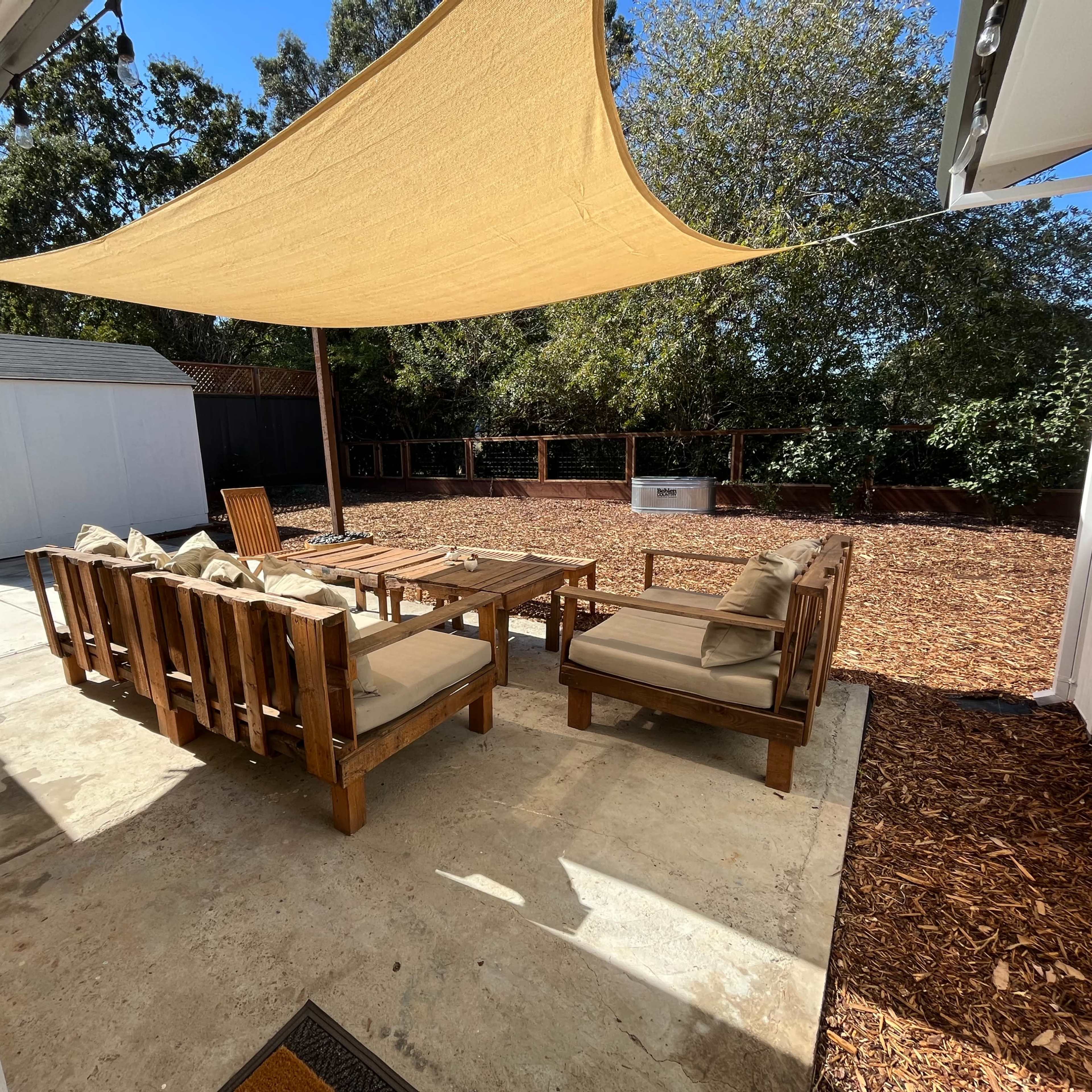 A shaded outdoor seating area with wooden furniture is set on a concrete patio surrounded by mulch.
