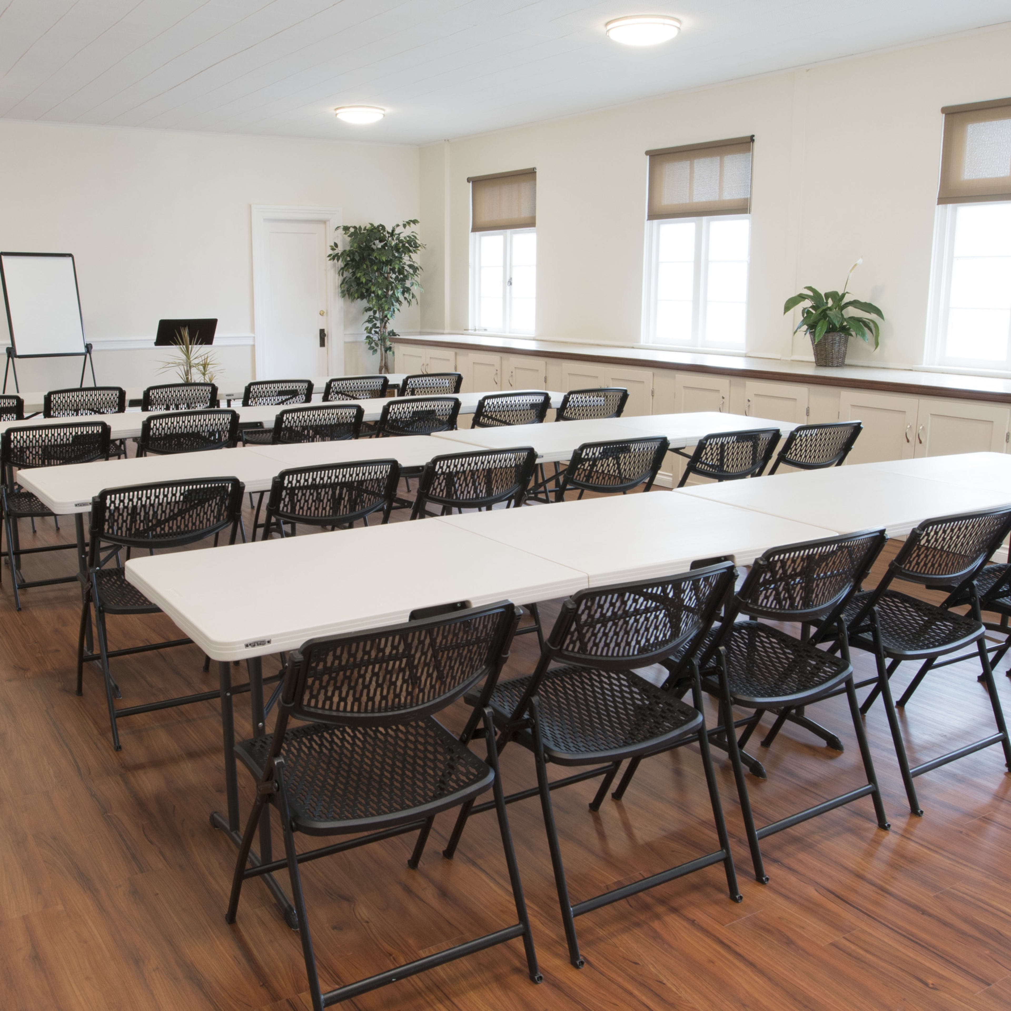 A conference room set up with neatly arranged folding tables and chairs, along with a flip chart and plants.
