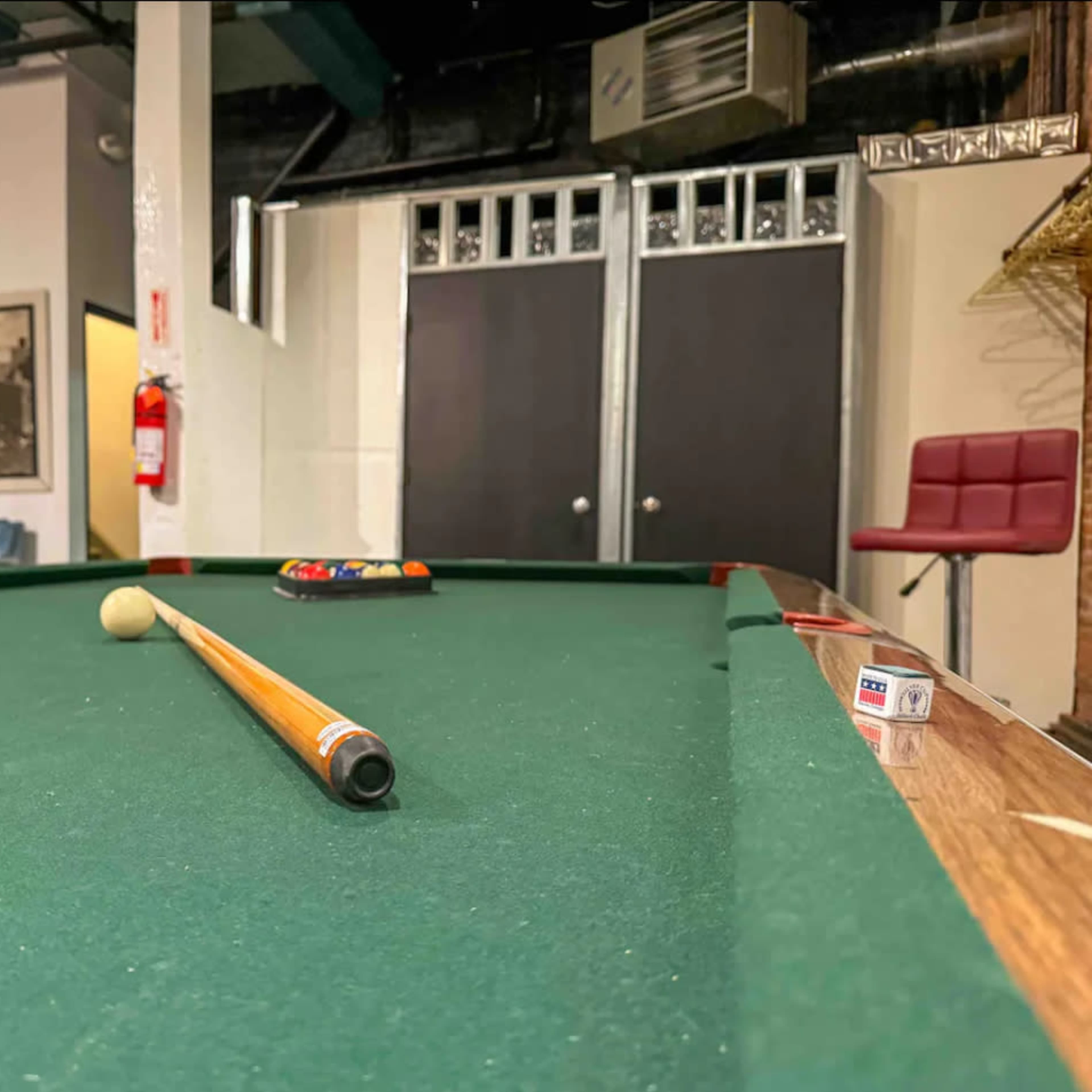 A pool table with a cue stick and a set of colored balls arranged in a rack is shown in a game room setting.