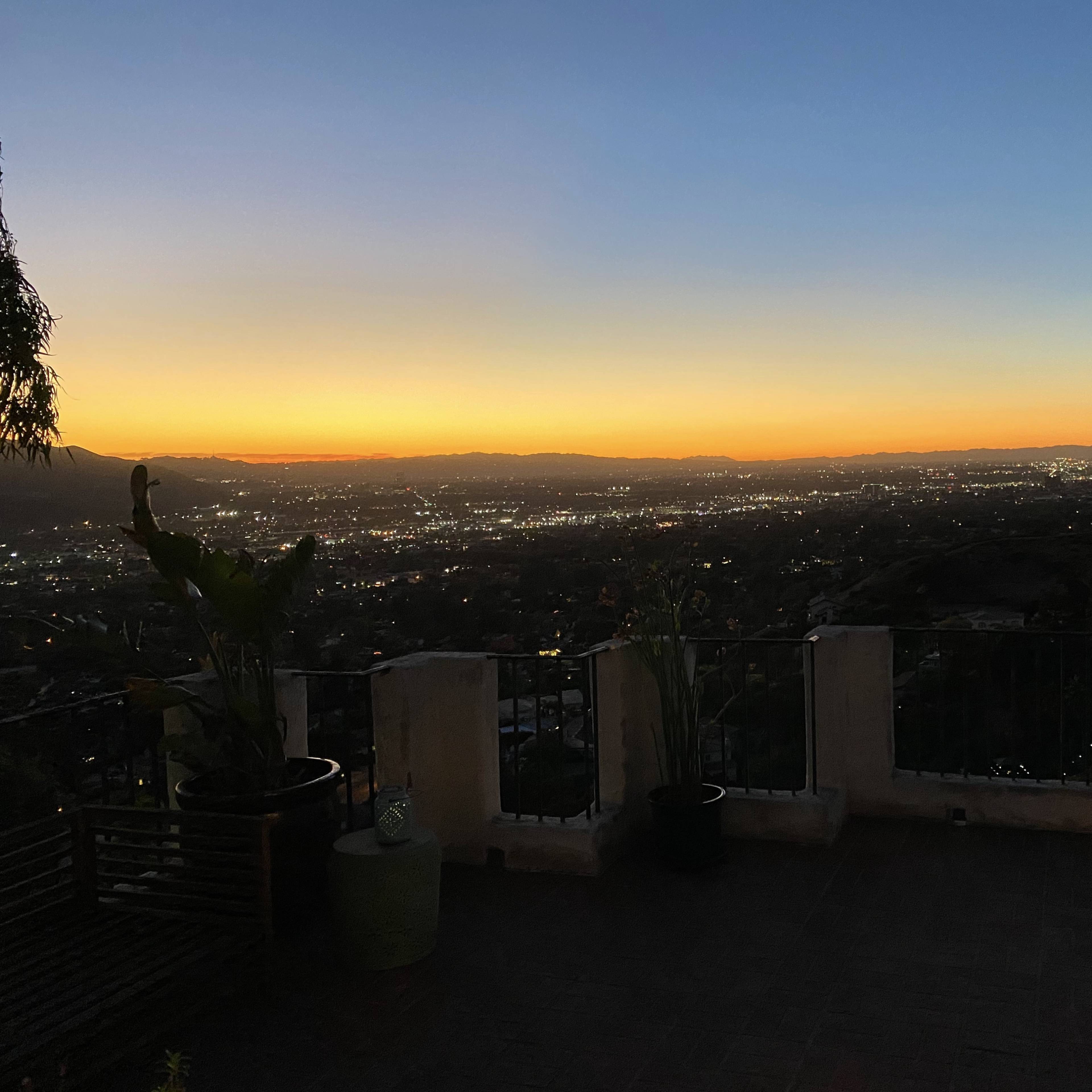 A sunset view over a city valley from a terrace, with distant lights scattered across the landscape.