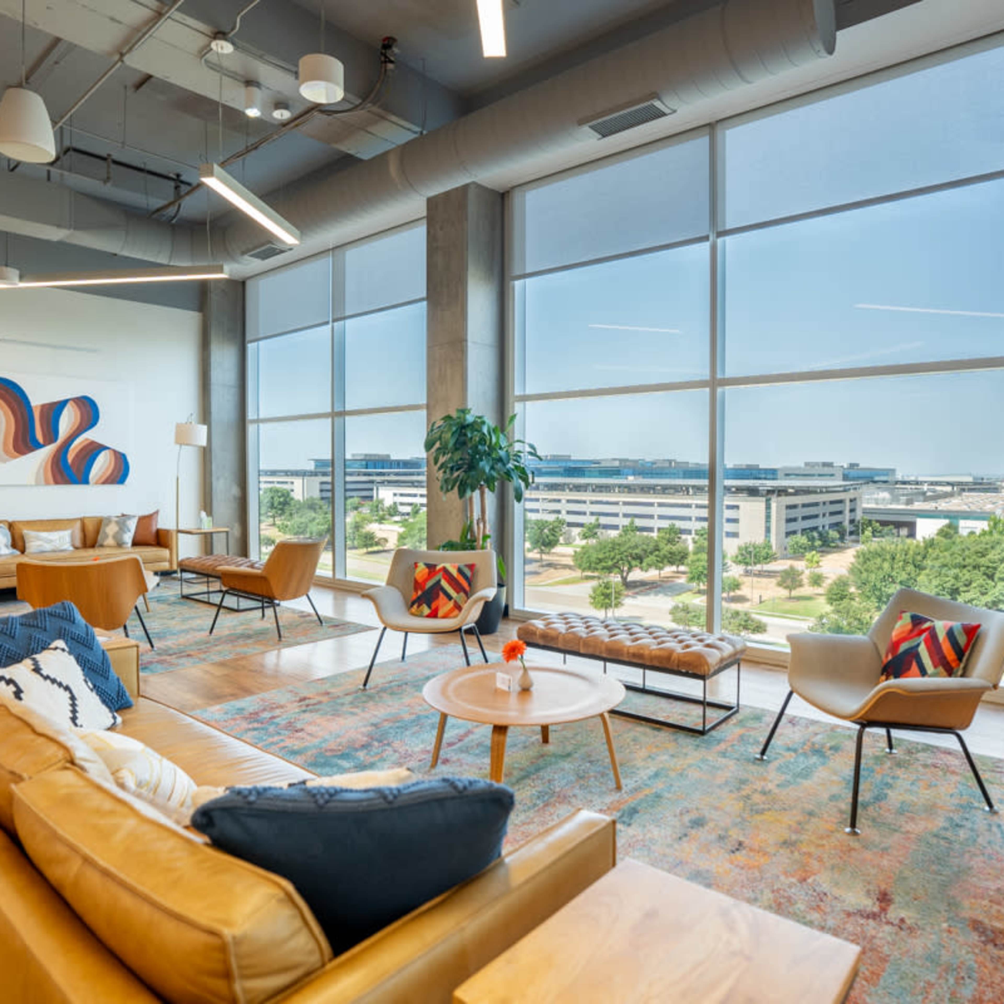 A modern office lounge area featuring a sofa, chairs, and large windows overlooking a landscaped outdoor space.