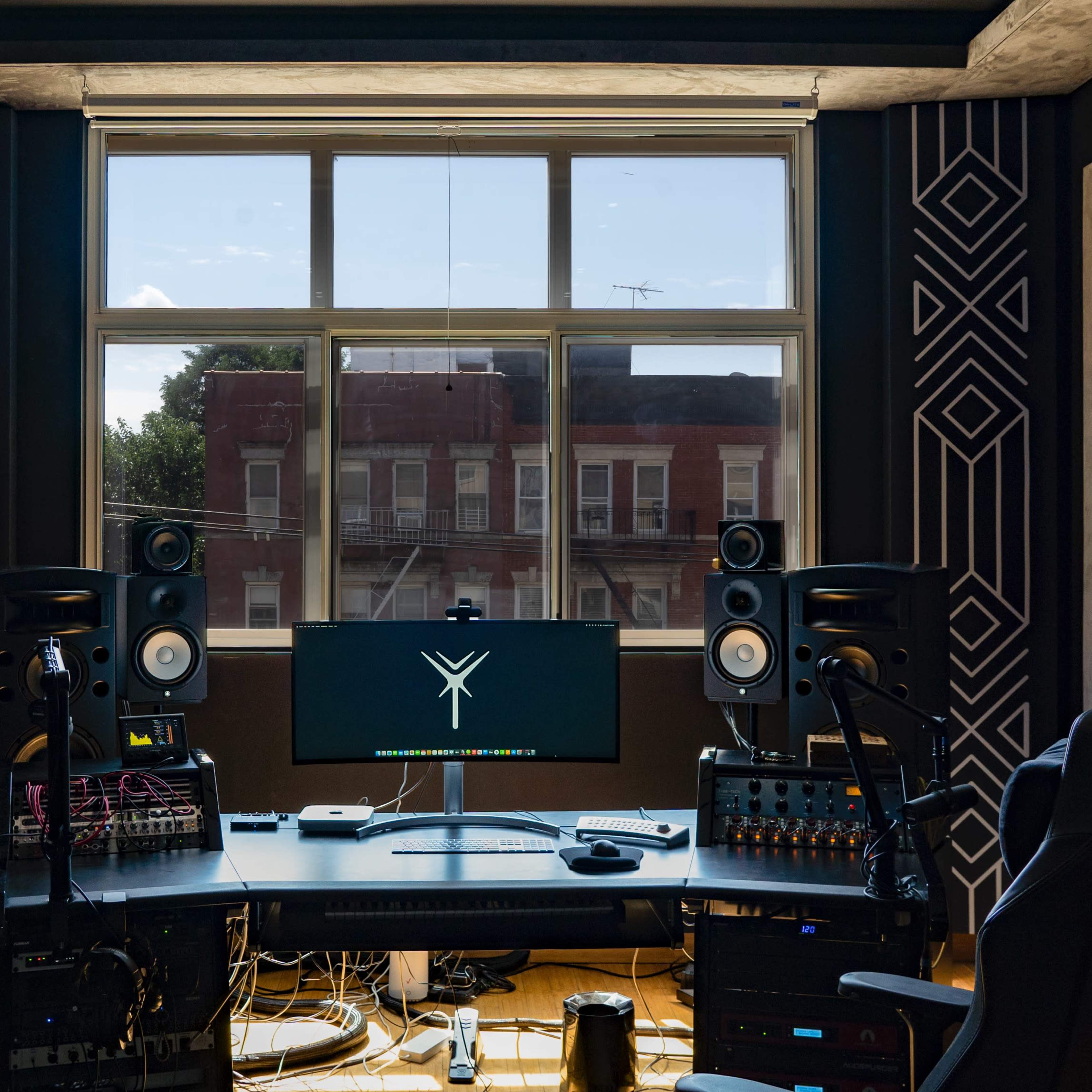 A music production studio features a large window with a view of a building across the street, surrounded by acoustic panels, a central desk with a computer monitor, speakers, and various audio equipment.