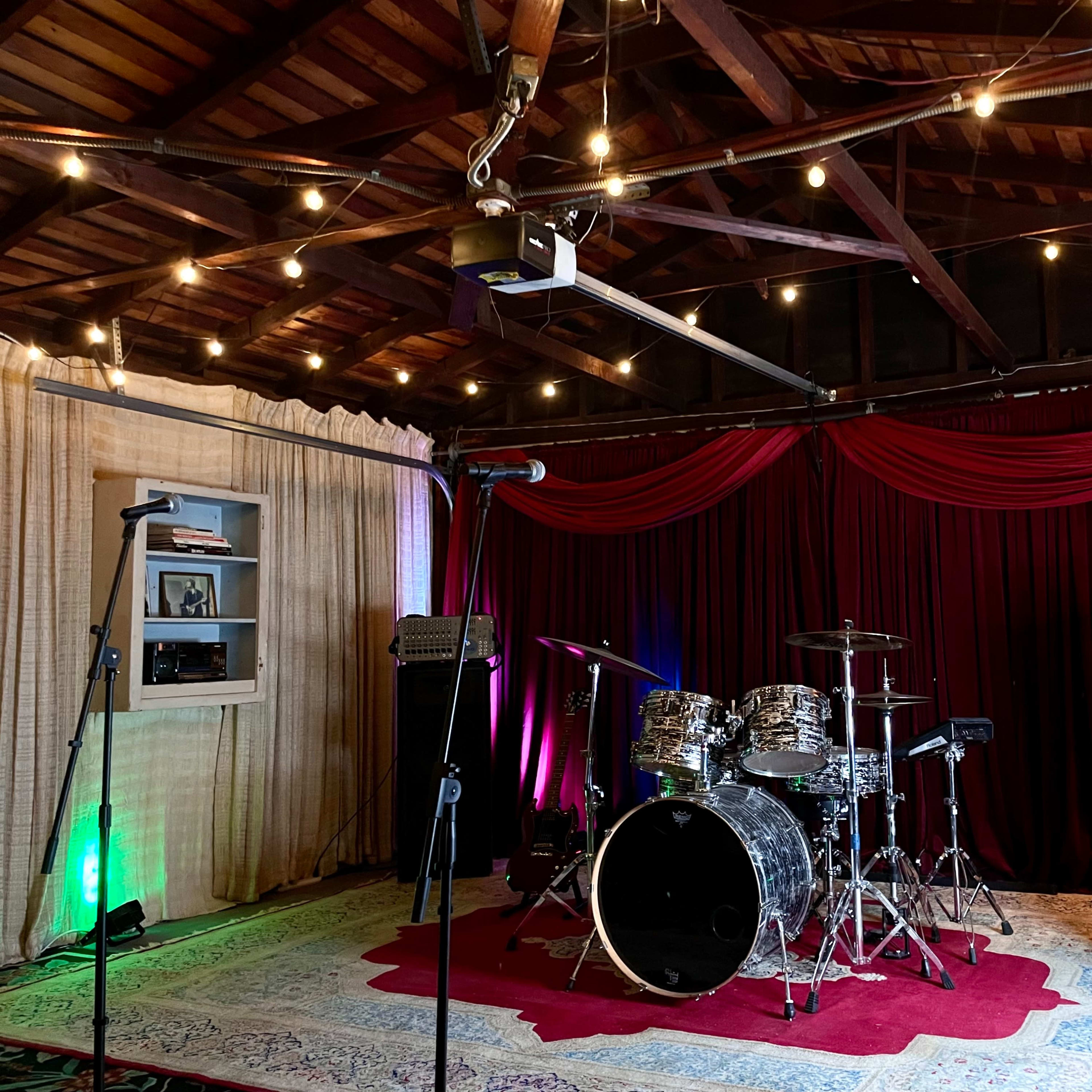 A music rehearsal space with a drum set, amplifiers, a microphone stand, and soft lighting from hanging lights and a lamp.