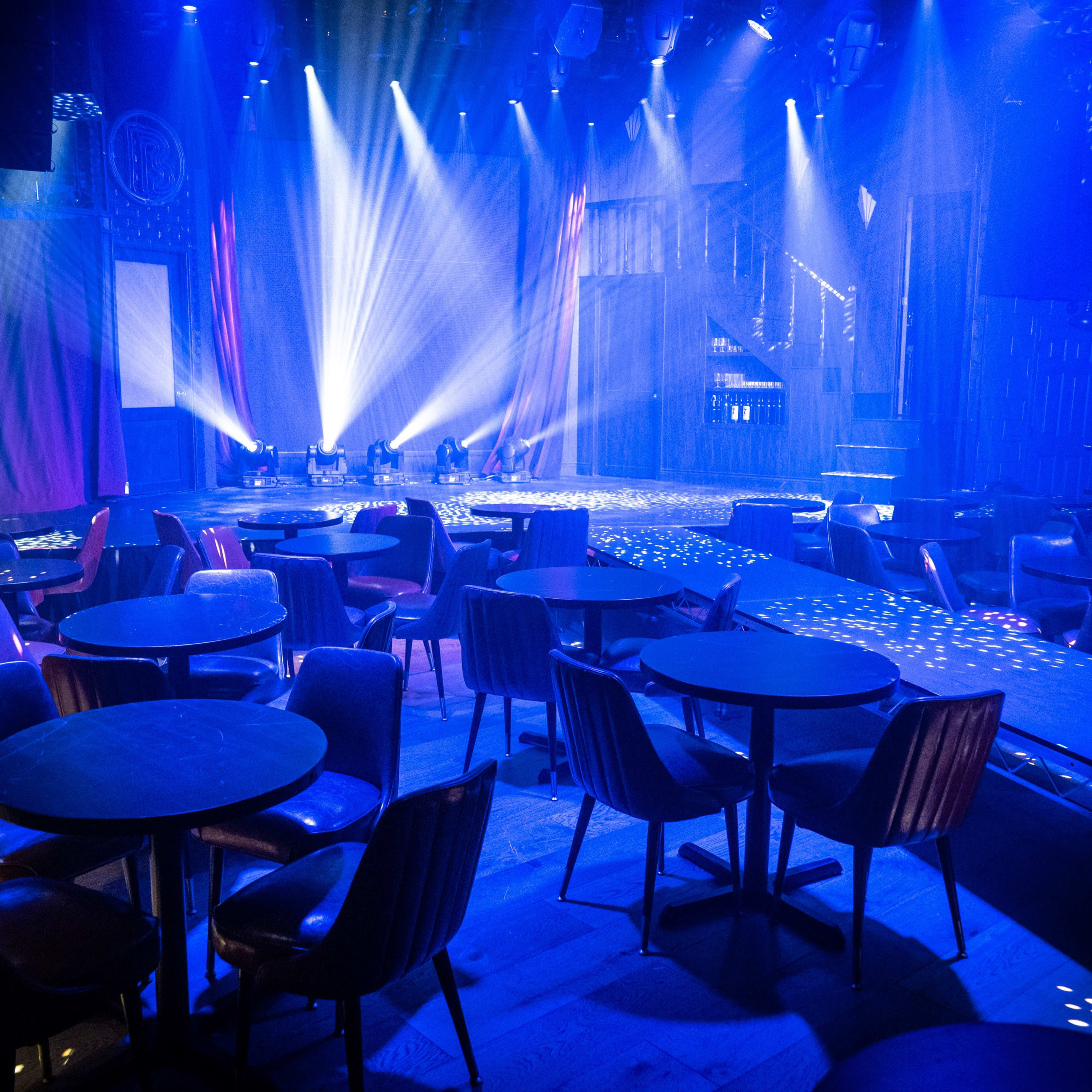 A dimly lit performance venue features circular tables and chairs with stage lights casting beams across the floor.