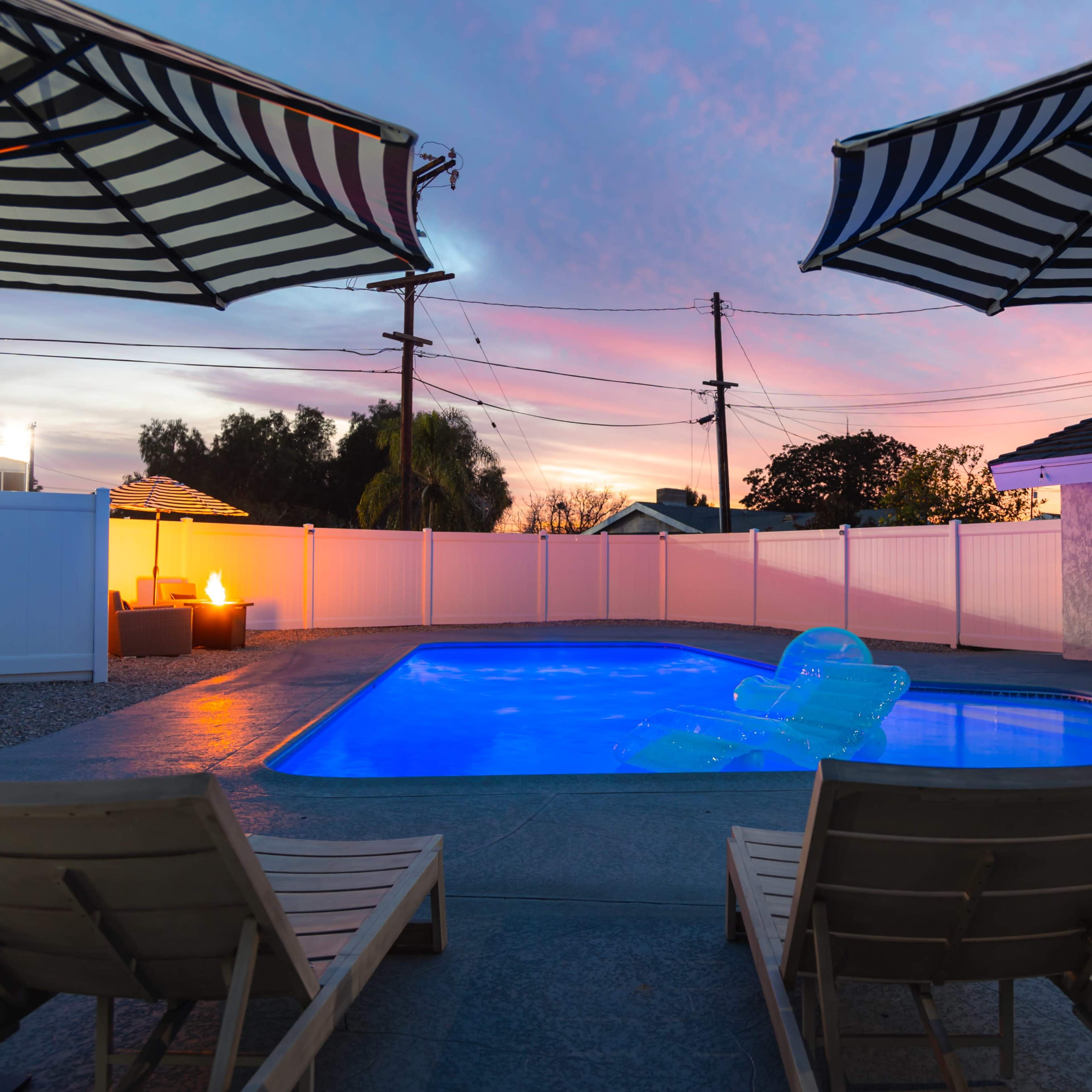 A swimming pool surrounded by two lounge chairs and striped umbrellas, with a sunset sky illuminating the scene.