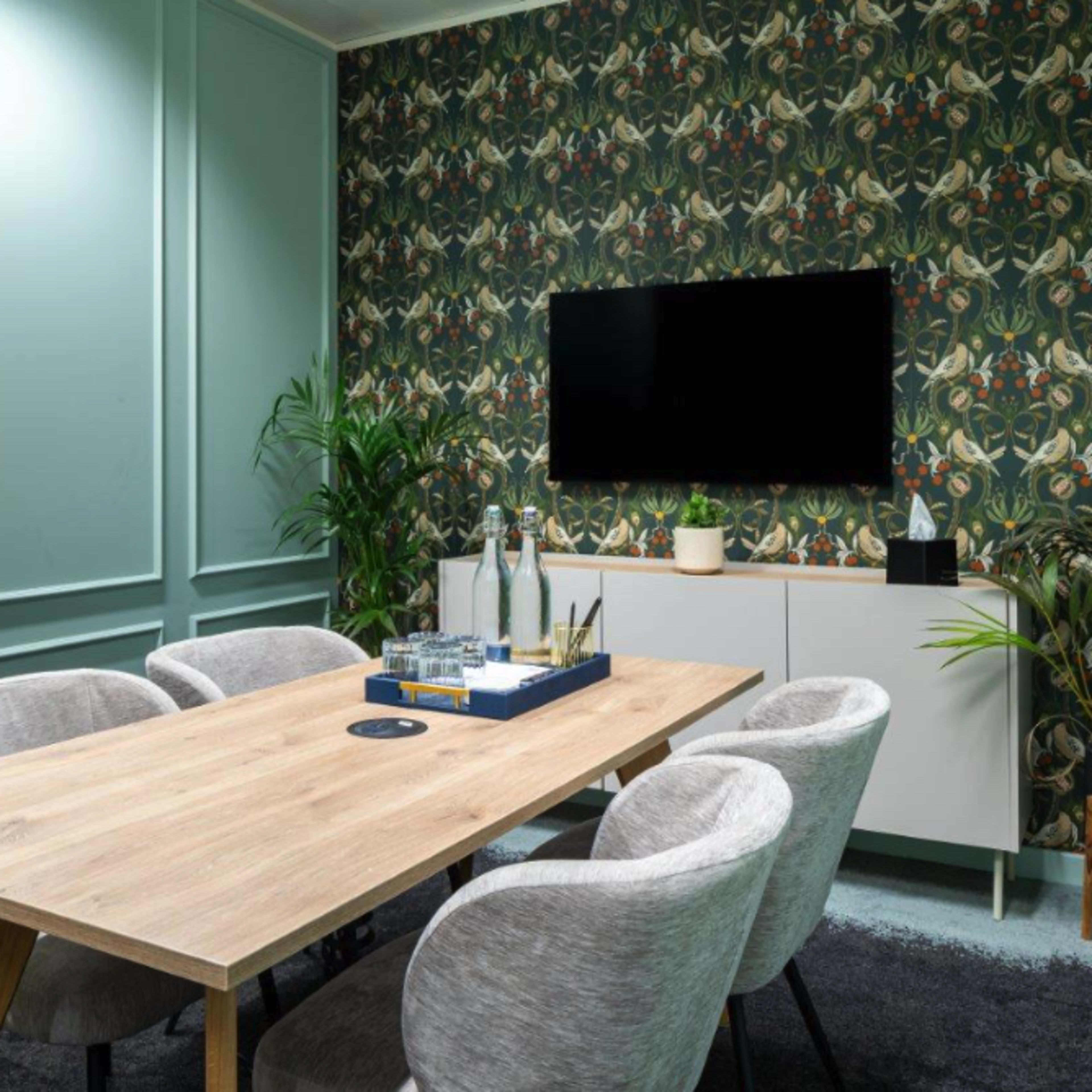 A modern conference room featuring a wooden table surrounded by four upholstered chairs, with a patterned wallpaper and a television mounted on the wall.