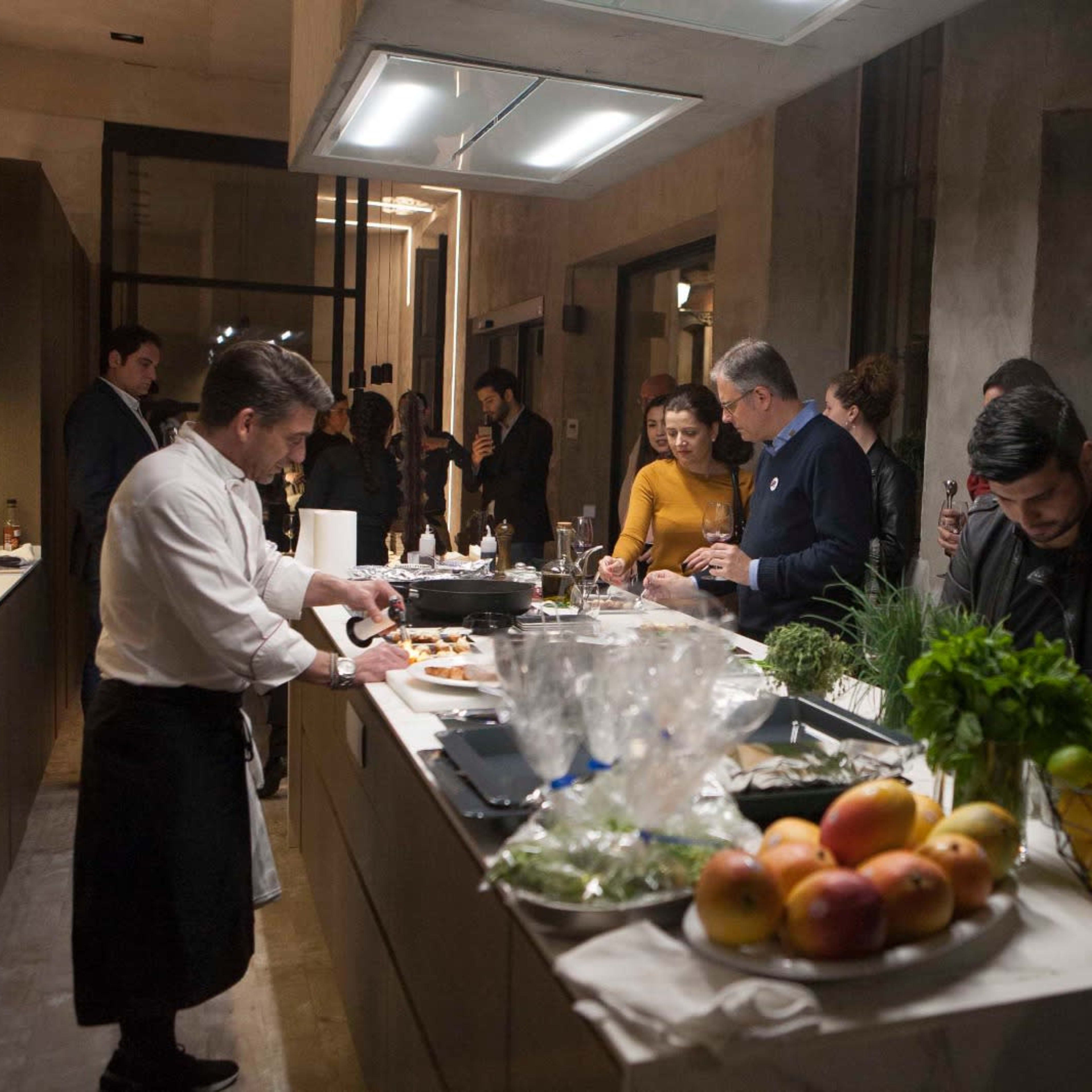 A group of people socializes in a modern kitchen while a chef prepares food on the counter, surrounded by fresh ingredients and fruits.