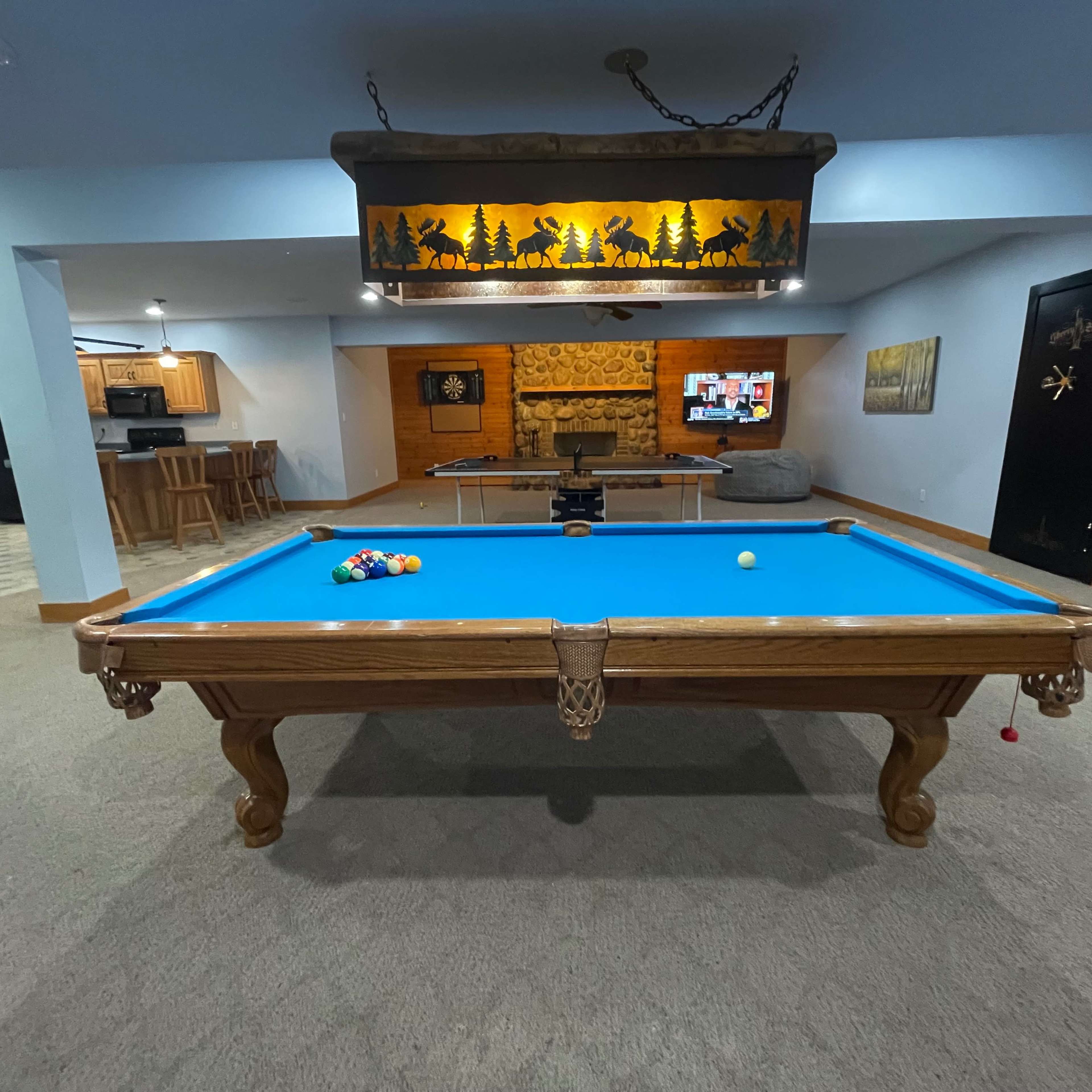 A well-lit game room features a blue pool table, a bar area in the background, and a television on the wall.