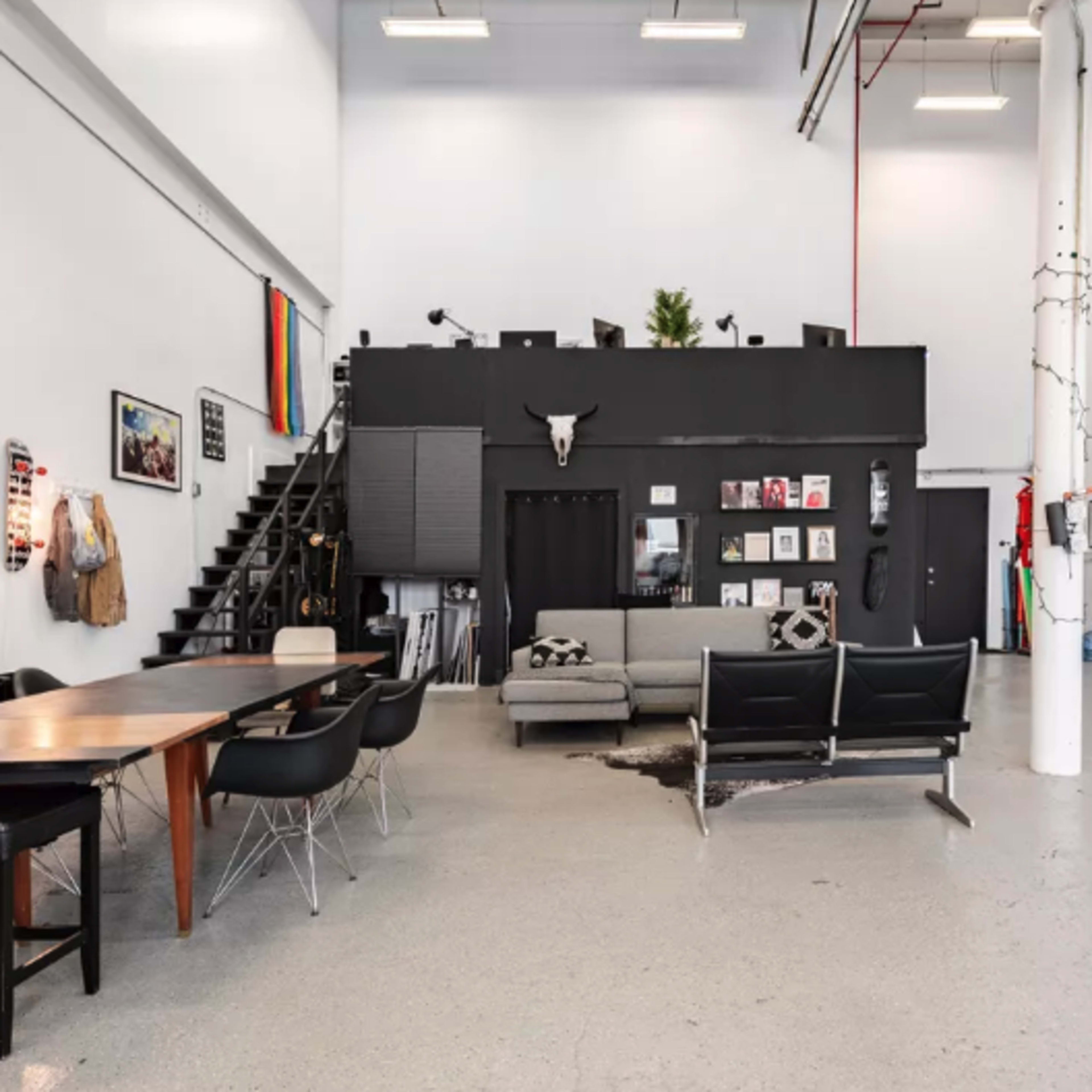 A spacious industrial-style room featuring a large dining table, black seating, an art-adorned wall, and a staircase leading to an upper level.