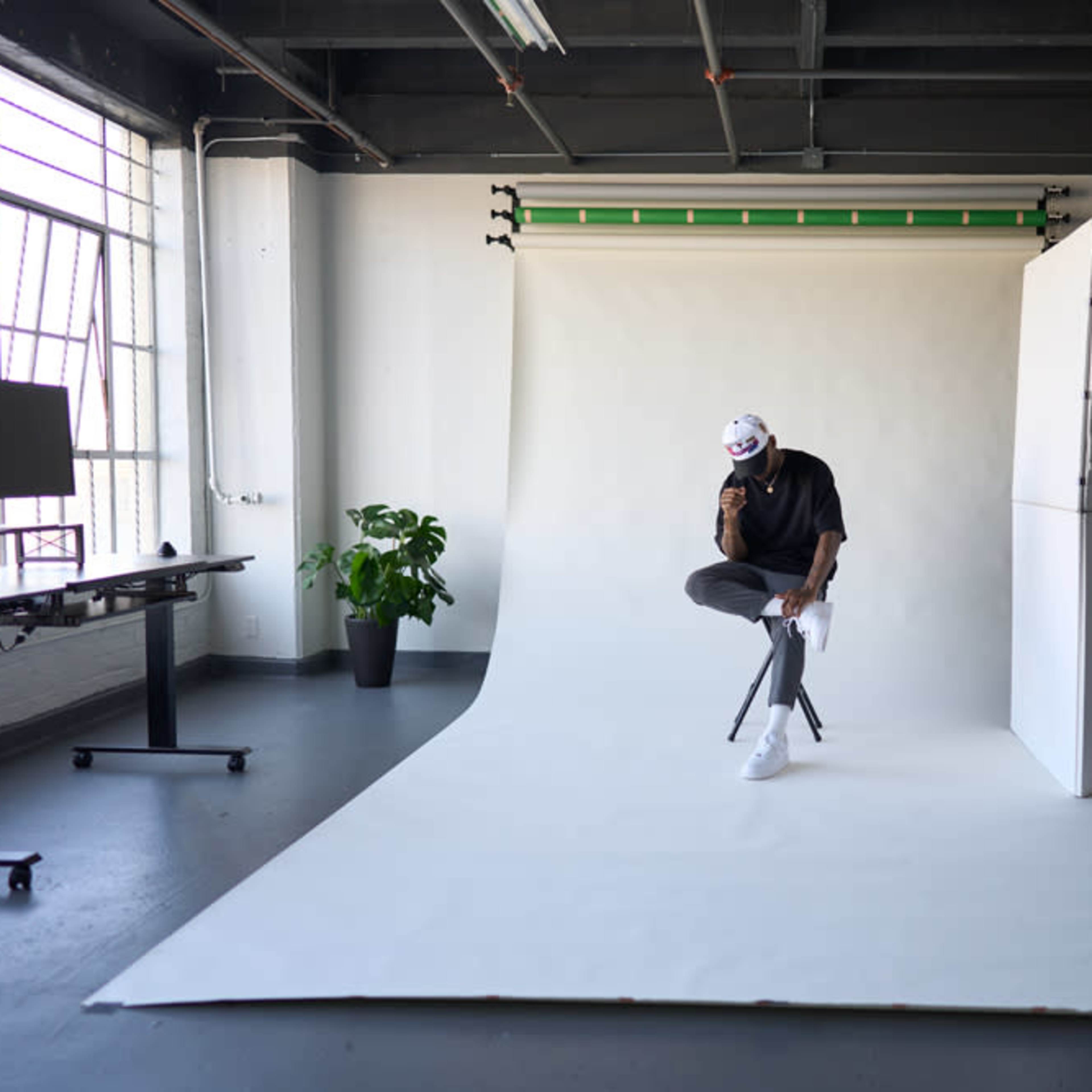 A person sits on a stool in a studio with a white backdrop and green lighting equipment.