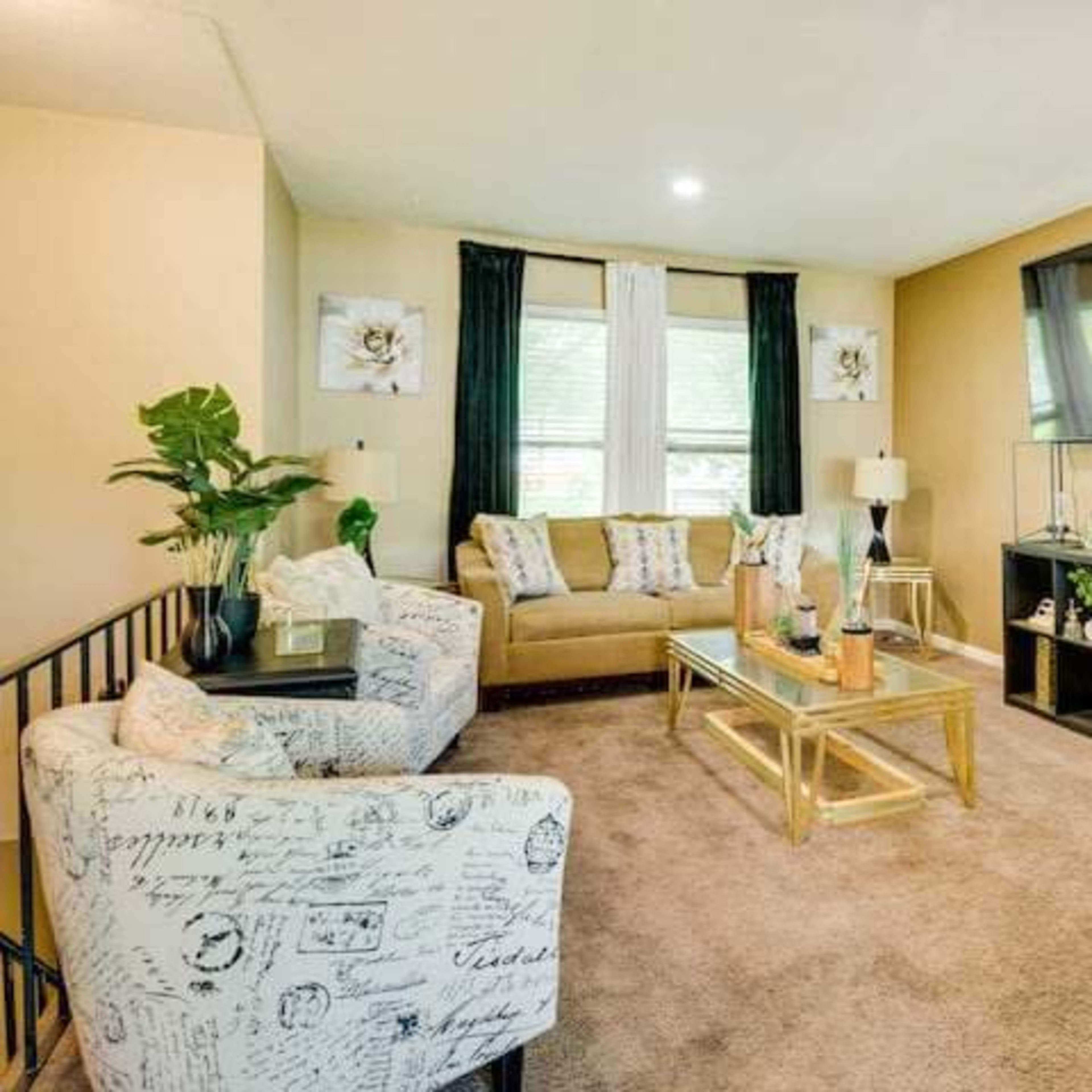 A modern living room featuring a tan sofa, two patterned chairs, a glass coffee table, and a large wall-mounted television.