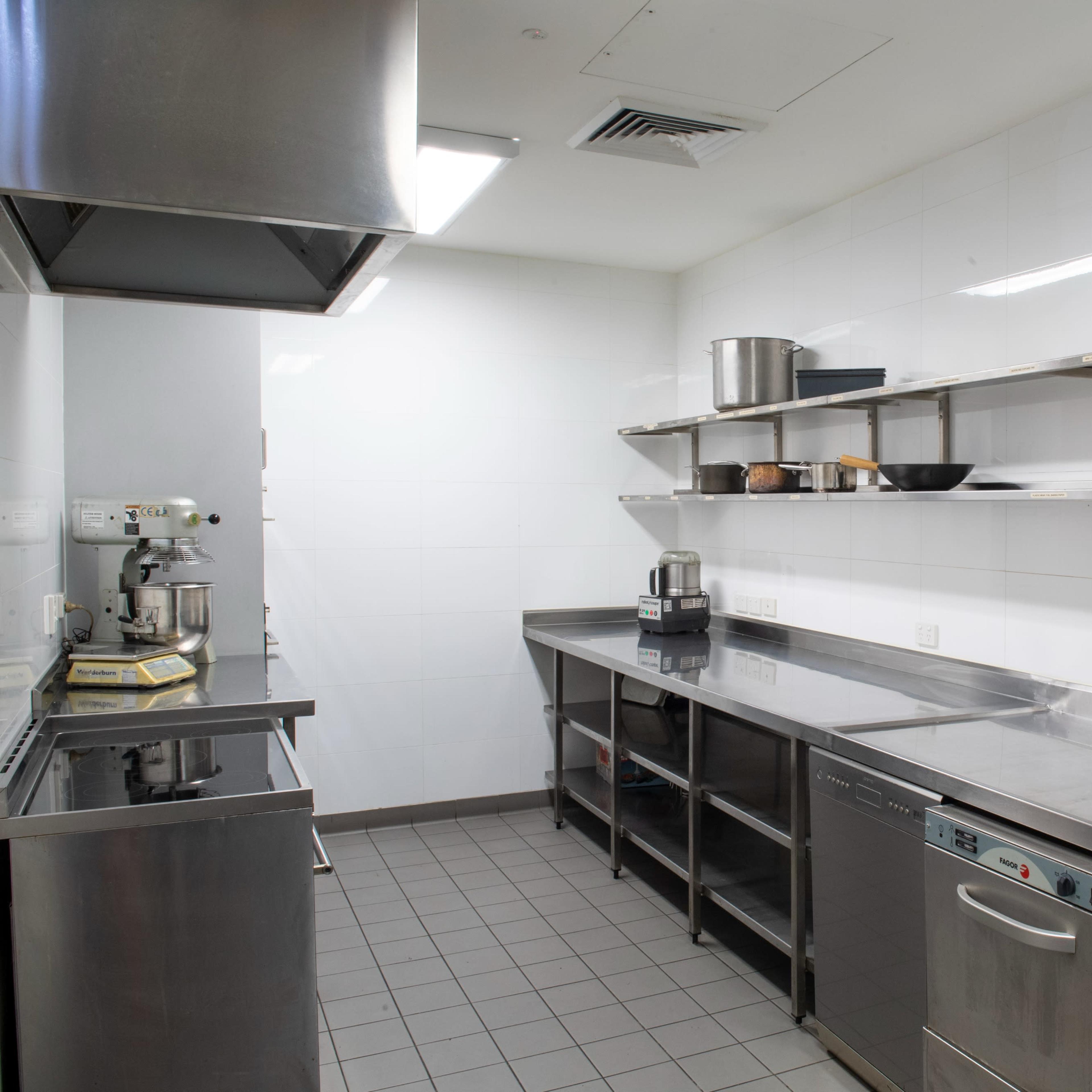 A commercial kitchen features stainless steel countertops, shelves with cooking utensils, and various appliances against white tiled walls.