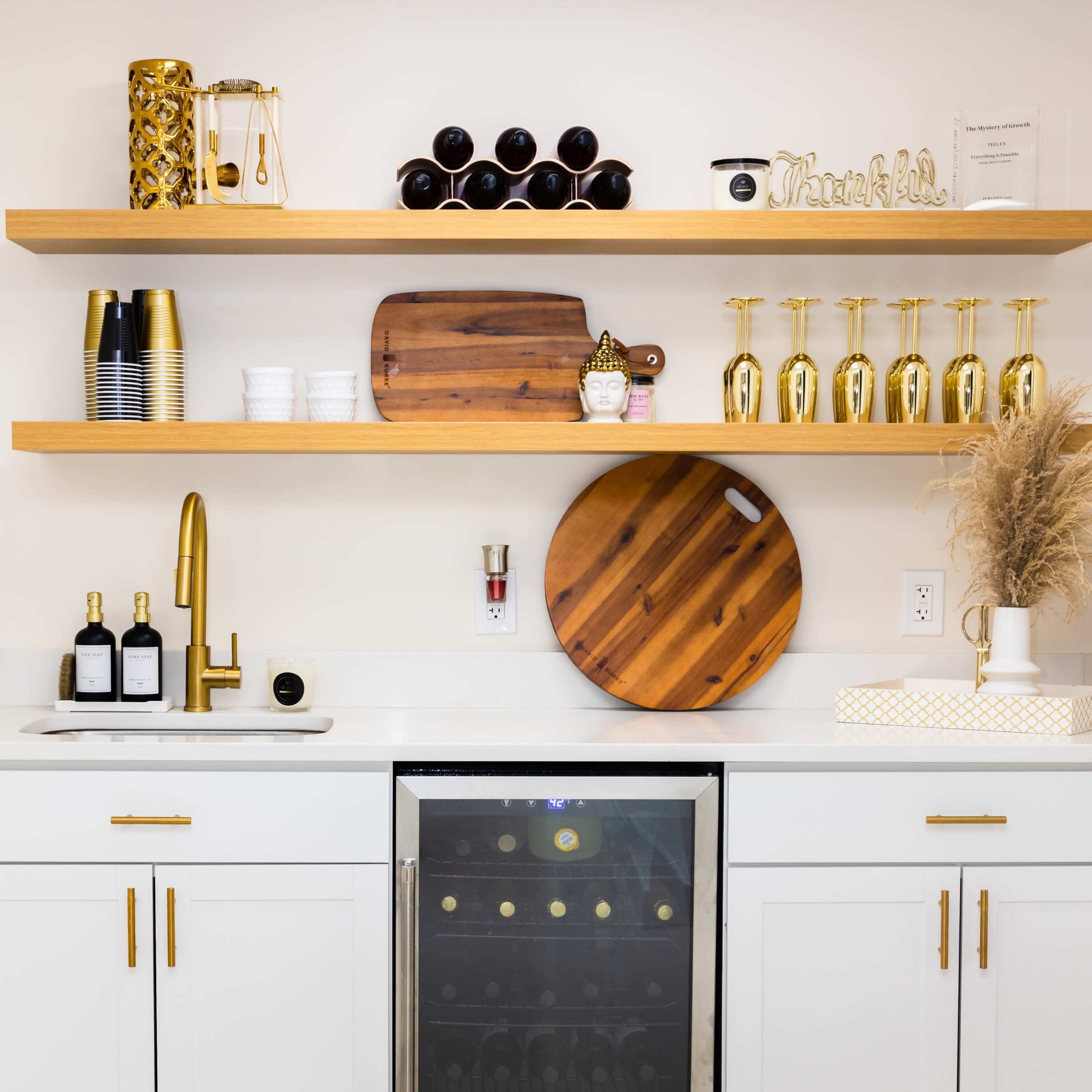 A modern kitchen bar area featuring white cabinets, two wooden shelves with decorative items, a wine cooler, and a gold faucet above a clean countertop.