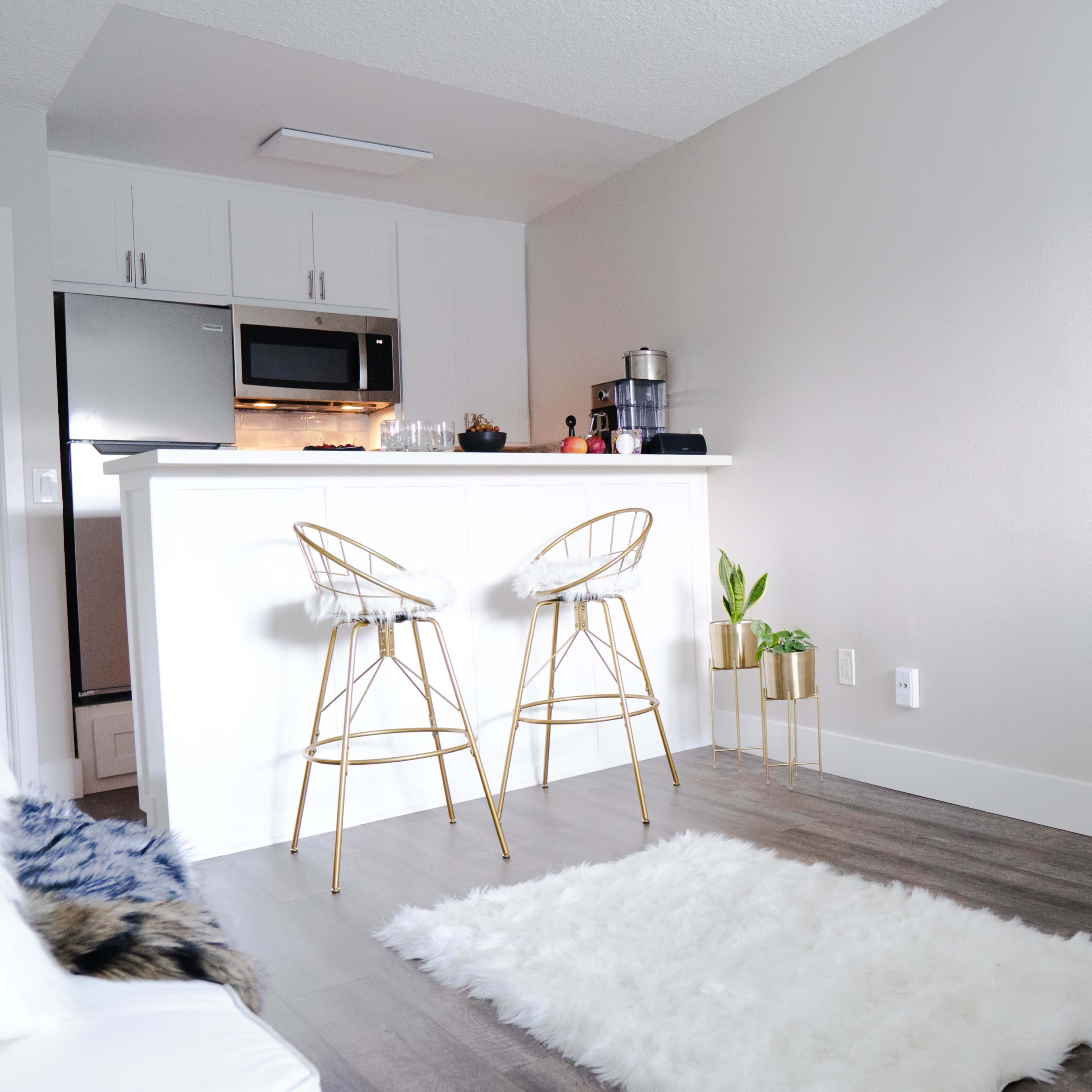 A modern living space featuring a small kitchen with a breakfast bar, two gold bar stools, a white rug on the floor, and a small table set with a chessboard.