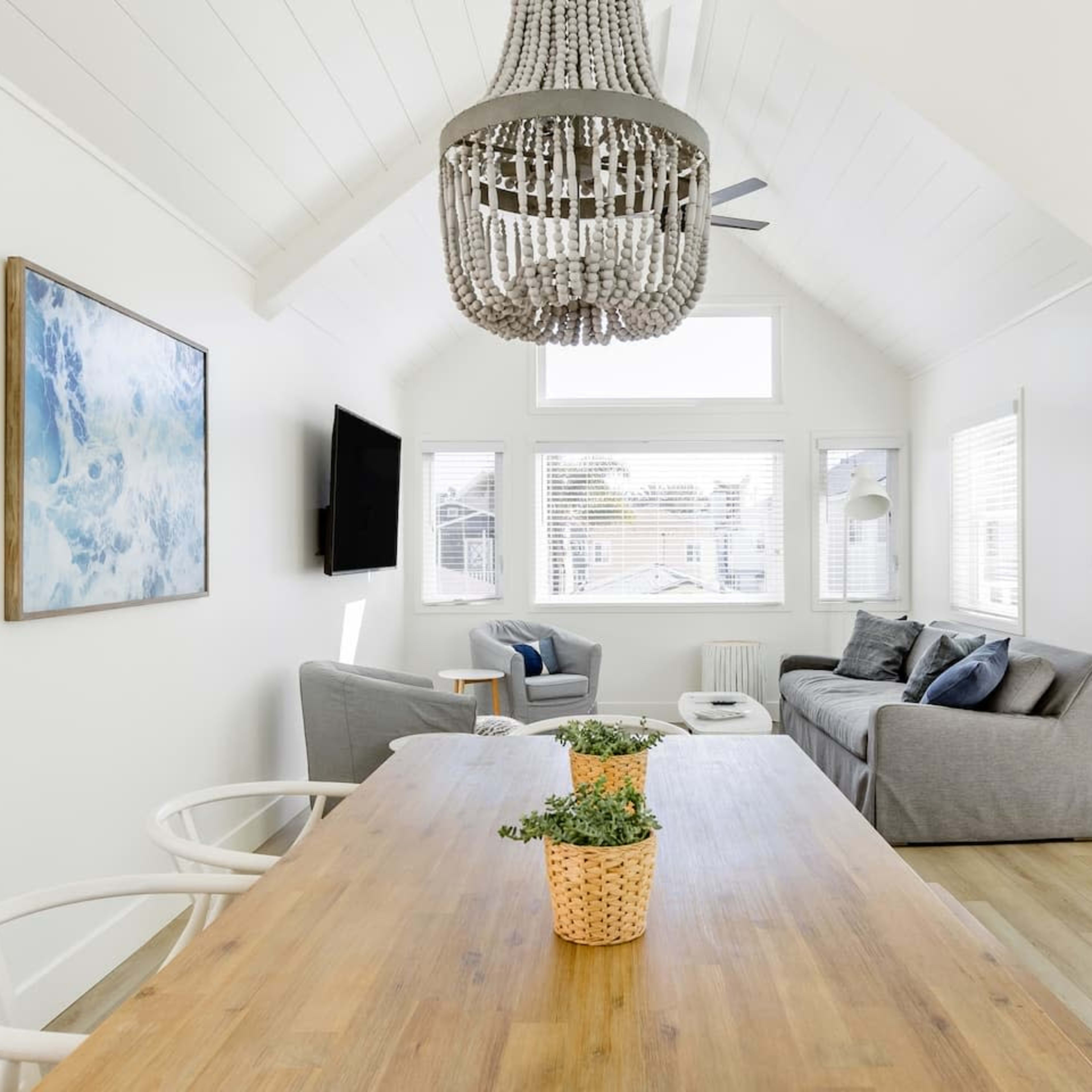 A bright, modern living space features a wooden dining table, a cozy seating area with gray sofas, large windows, and a decorative chandelier.
