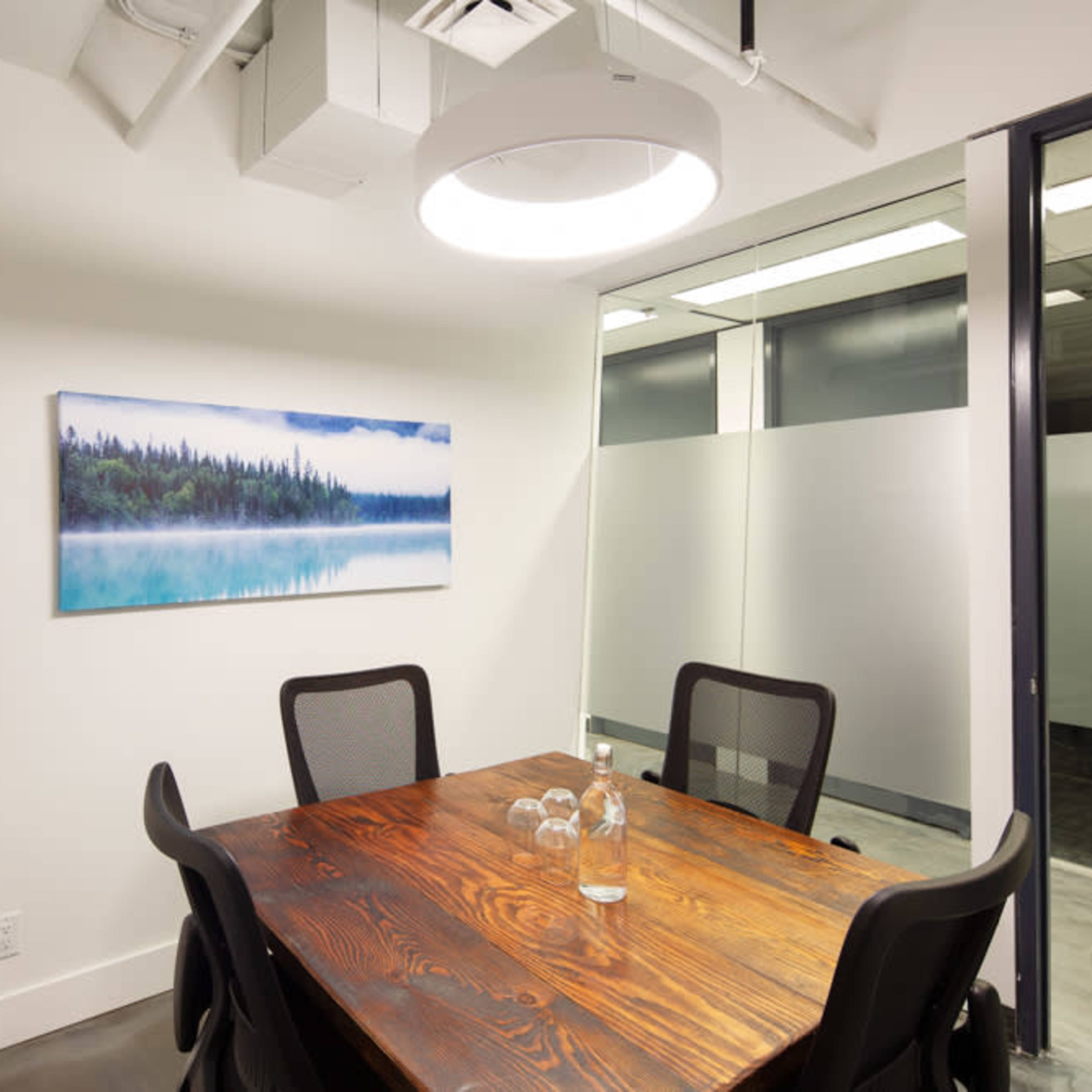 A small conference room features a wooden table surrounded by four black chairs, with a large window showcasing a view of a forested landscape painting.