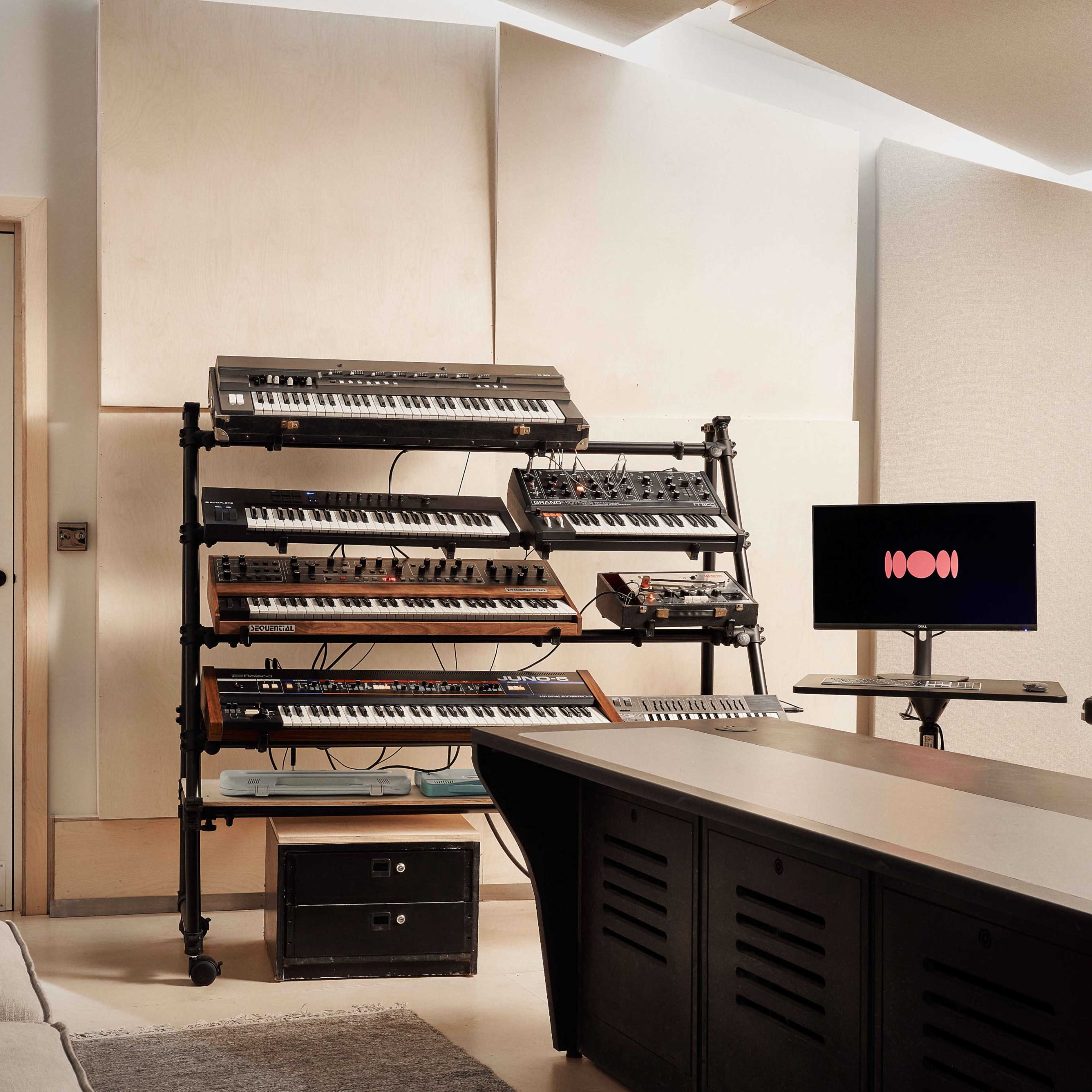 A spacious music studio features a drum set, keyboards, and multiple microphones arranged on stands, with a large window and natural light illuminating the room.