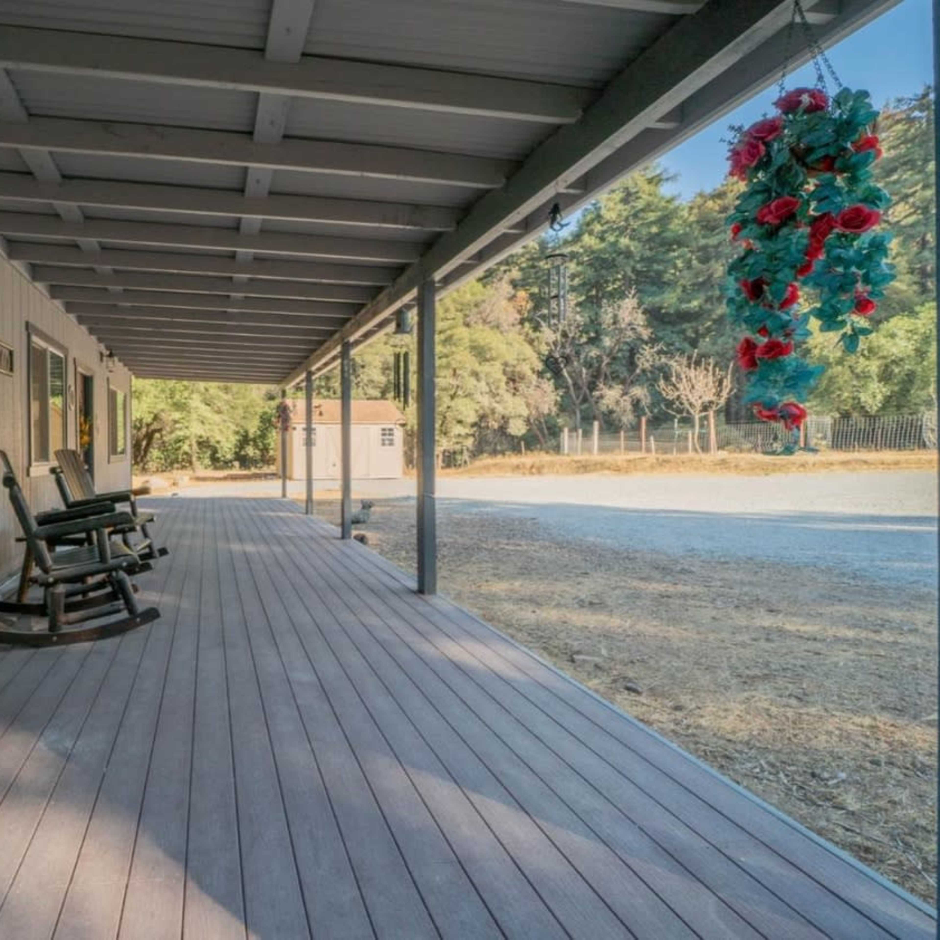 A porch with wooden flooring, featuring rockers and a hanging flower arrangement, alongside a gravel driveway and trees in the background.