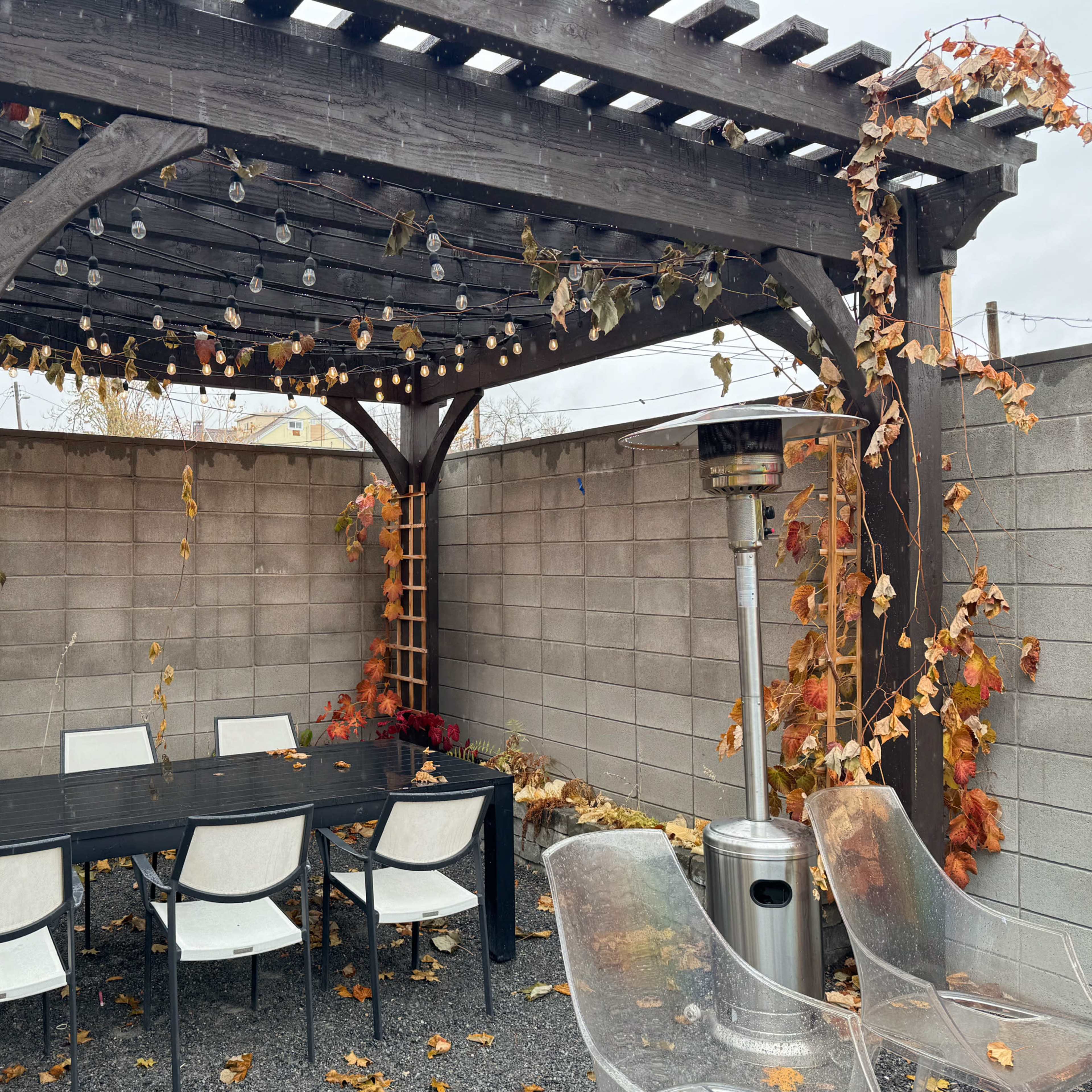 A wooden pergola with string lights surrounds a rectangular dining table and chairs, situated in a gravel courtyard with a grey brick wall.