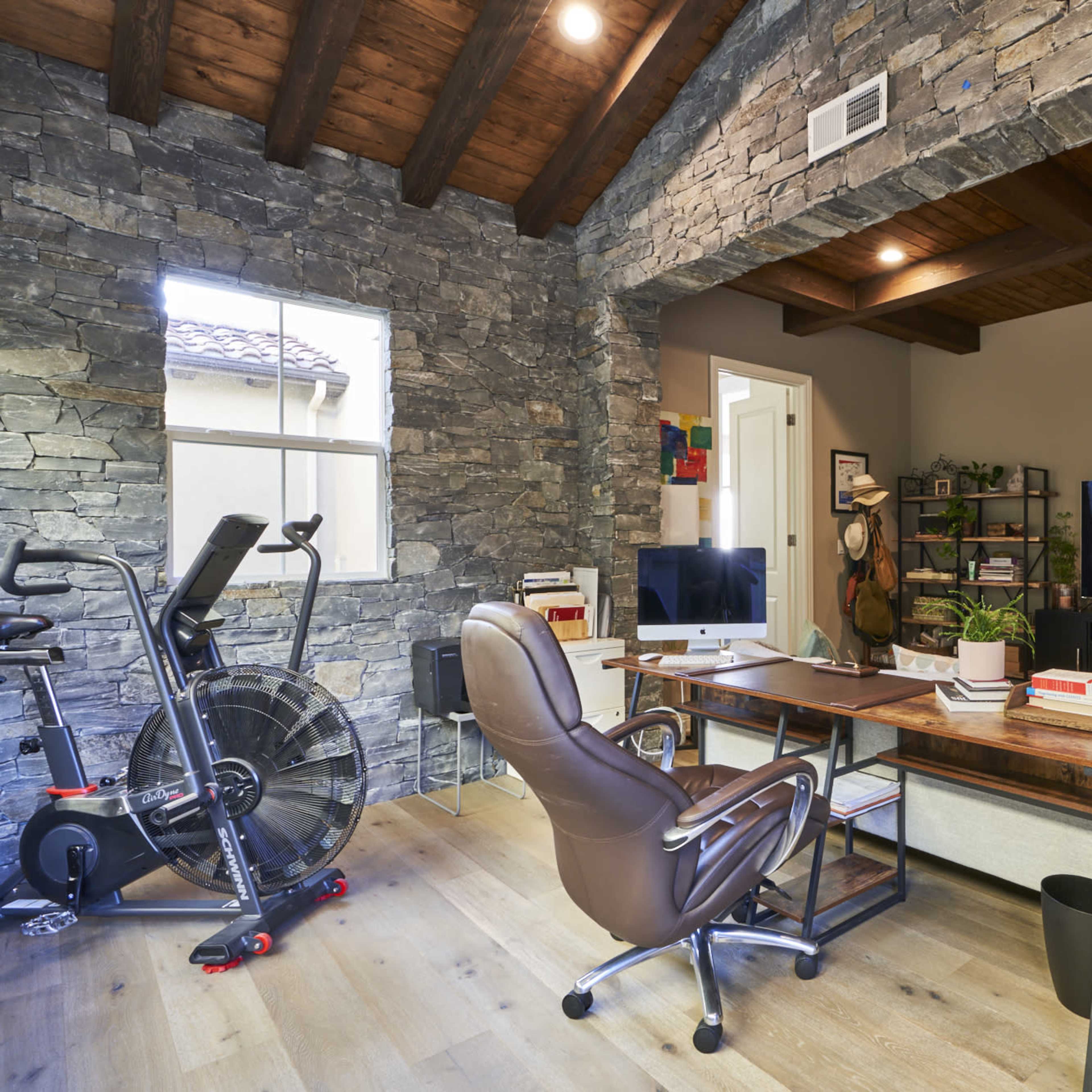 A modern home office with a wooden desk, an ergonomic chair, a computer, a bookshelf filled with plants and books, and a stationary exercise bike in a room featuring stone walls and wooden beams.