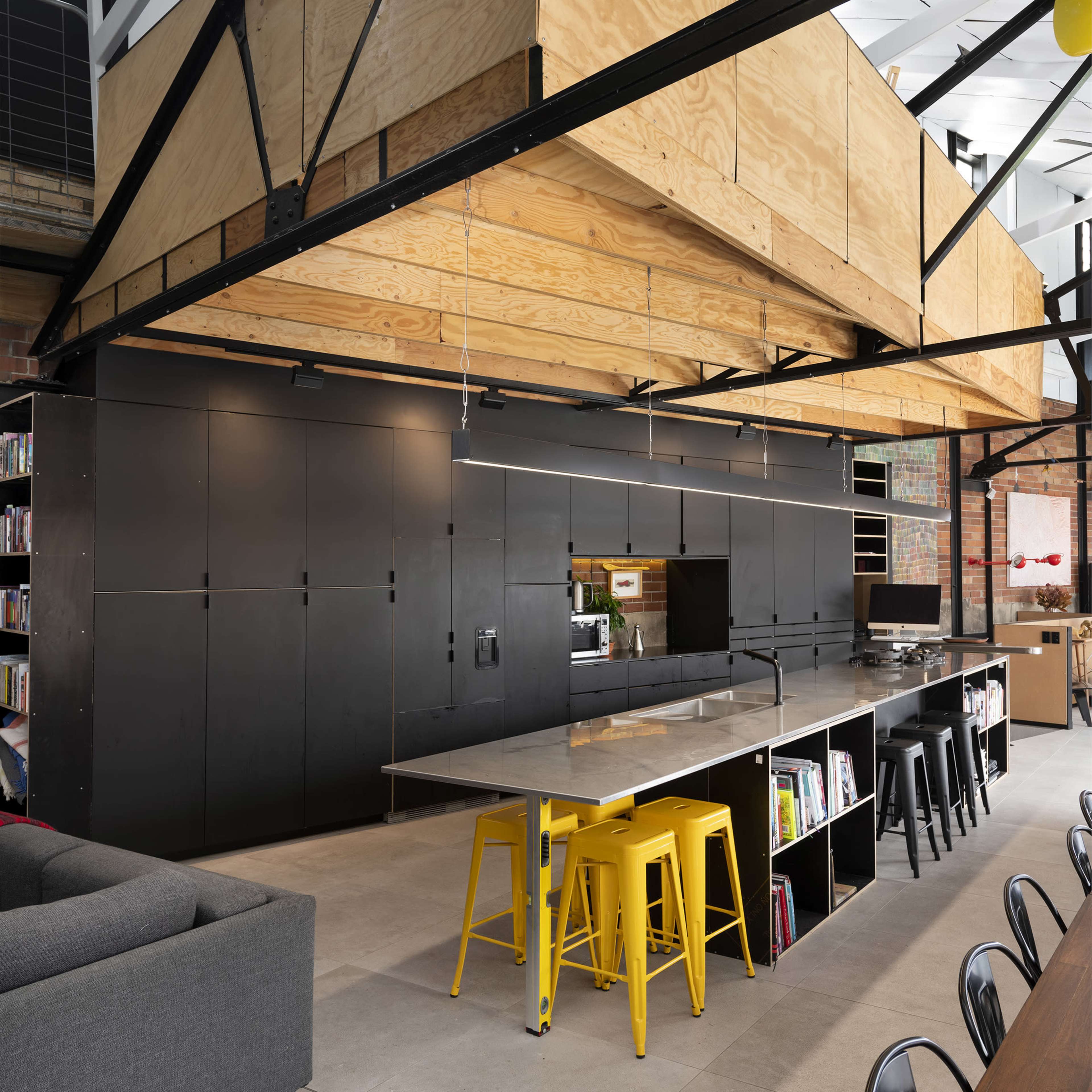 A modern kitchen and dining area featuring a large wooden structure overhead, with black cabinetry and yellow stools at a long gray table.