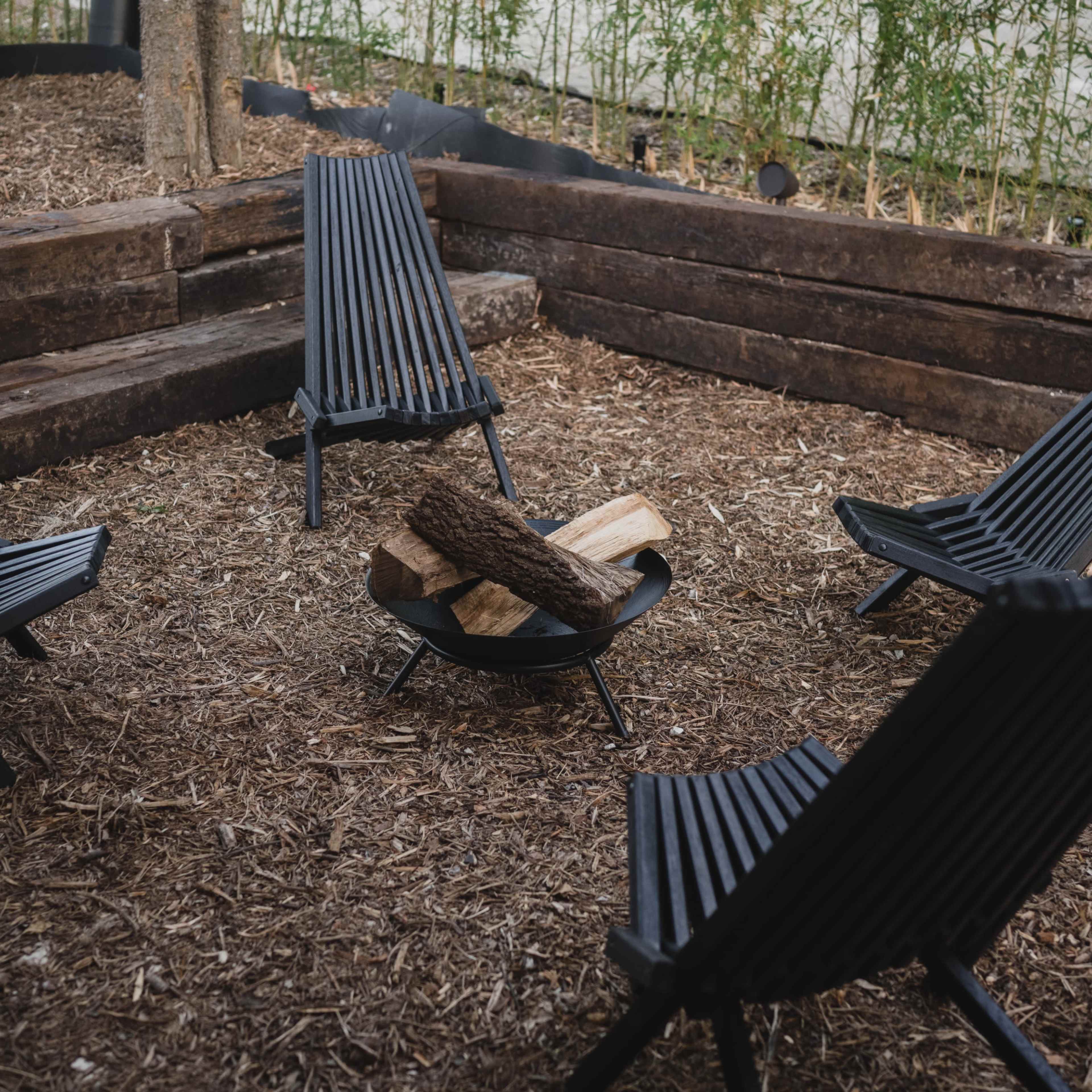 A circular seating area with four black chairs surrounding a fire pit containing logs, all set on a bed of mulch in a garden space.