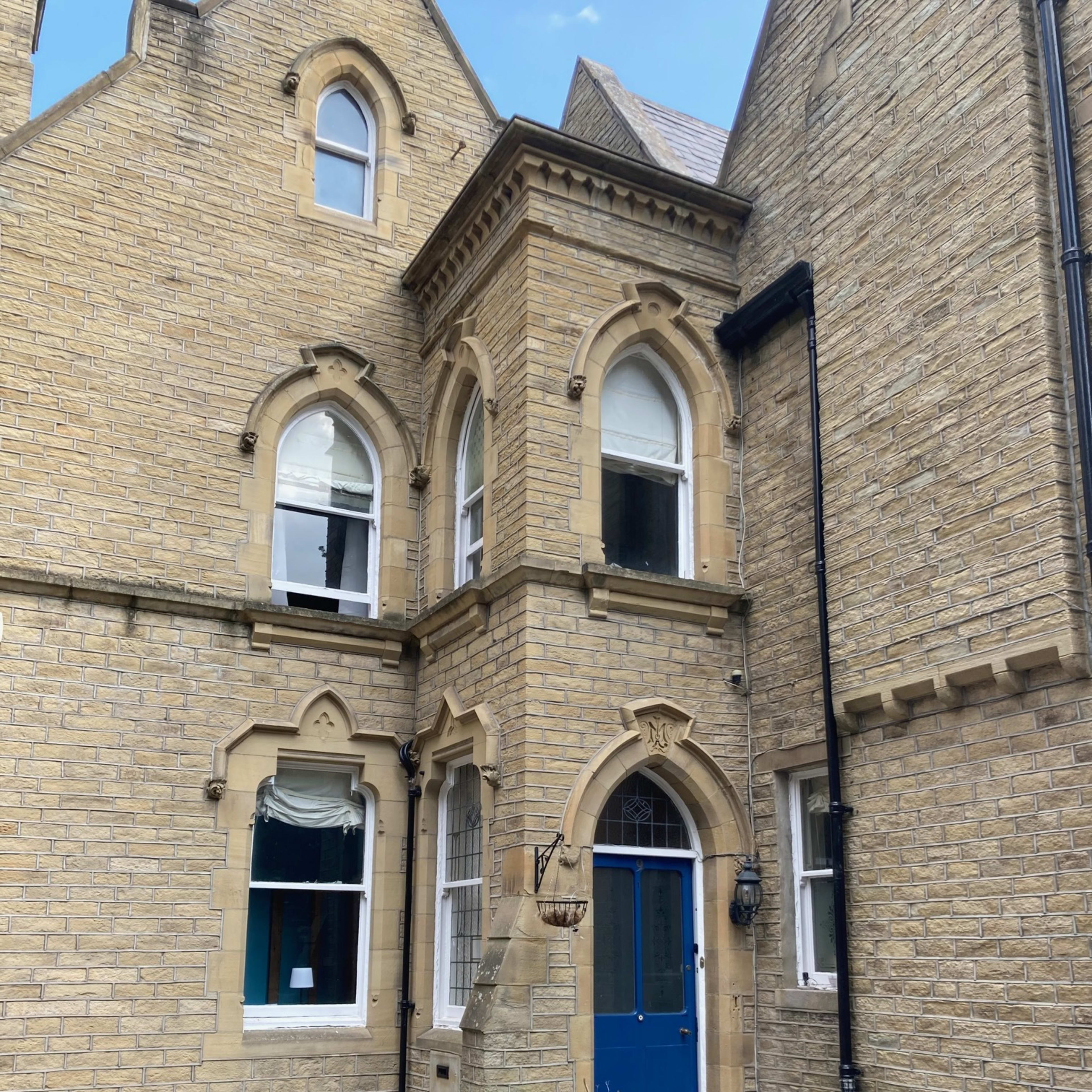 A stone building with arched windows, featuring a blue door at the entrance and a small garden area in front.