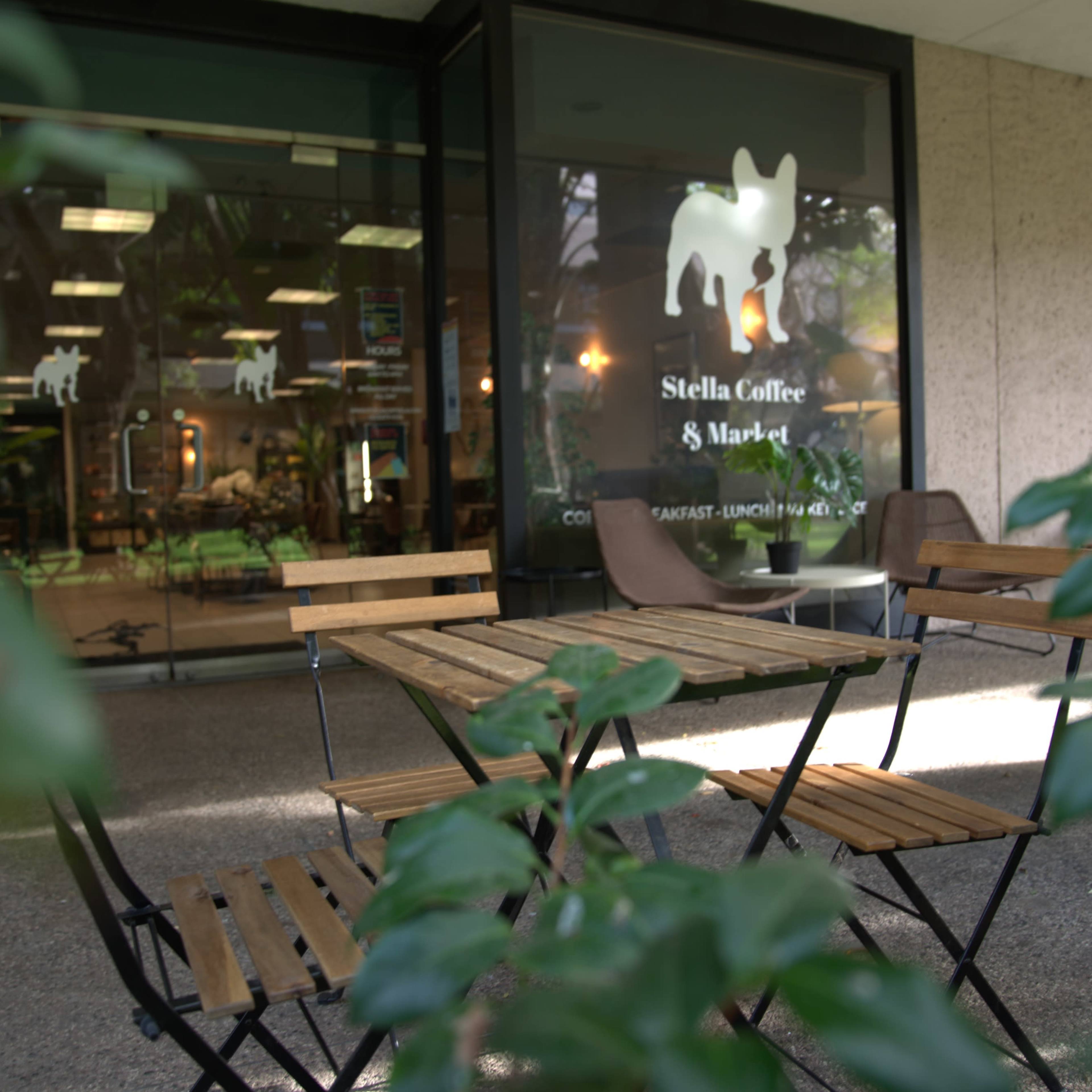 A small outdoor seating area with wooden tables and chairs is positioned in front of a café named "Stella Coffee & Market," visible through glass doors.