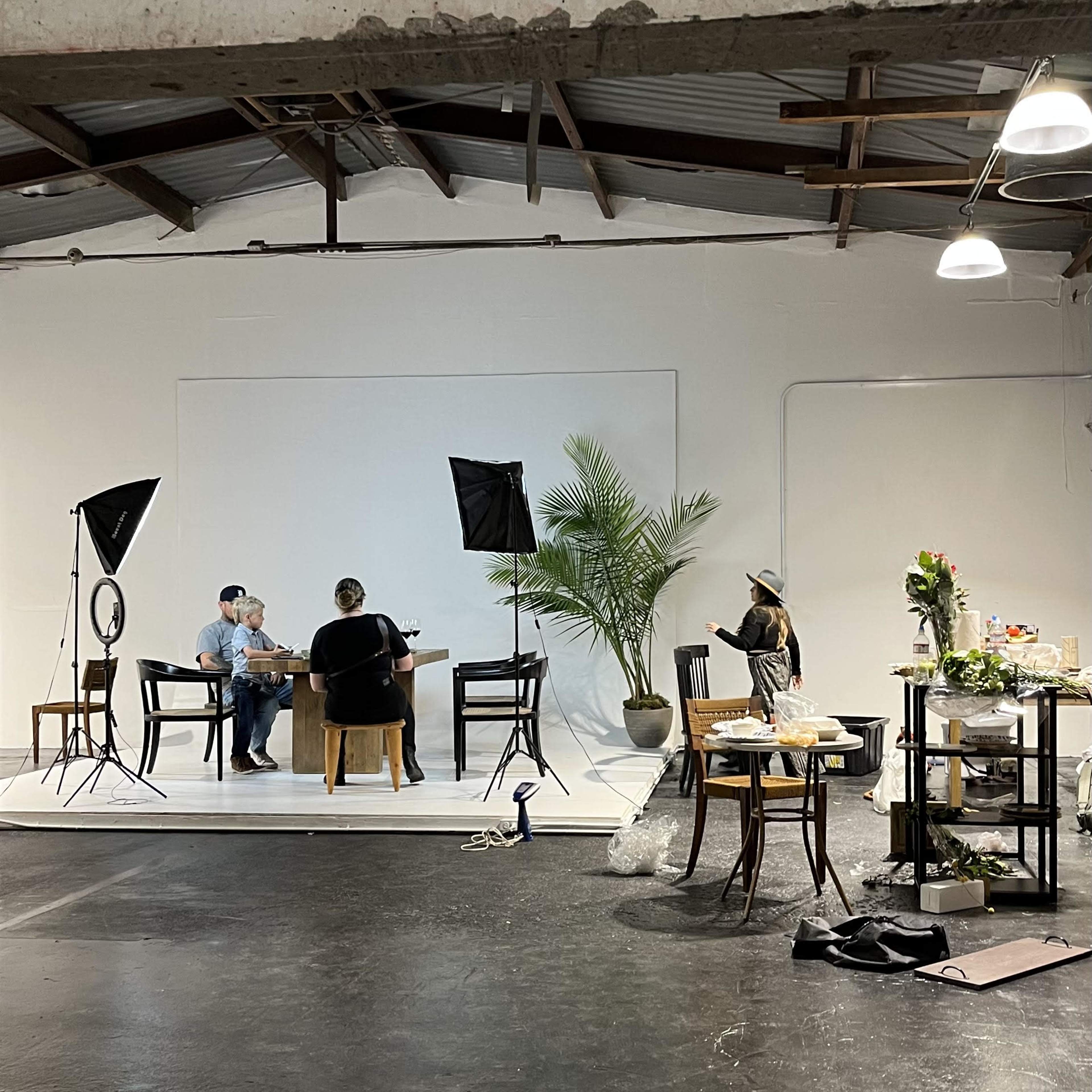 A studio setup features two people seated at a table in front of a plain backdrop, with camera equipment and props arranged around them.
