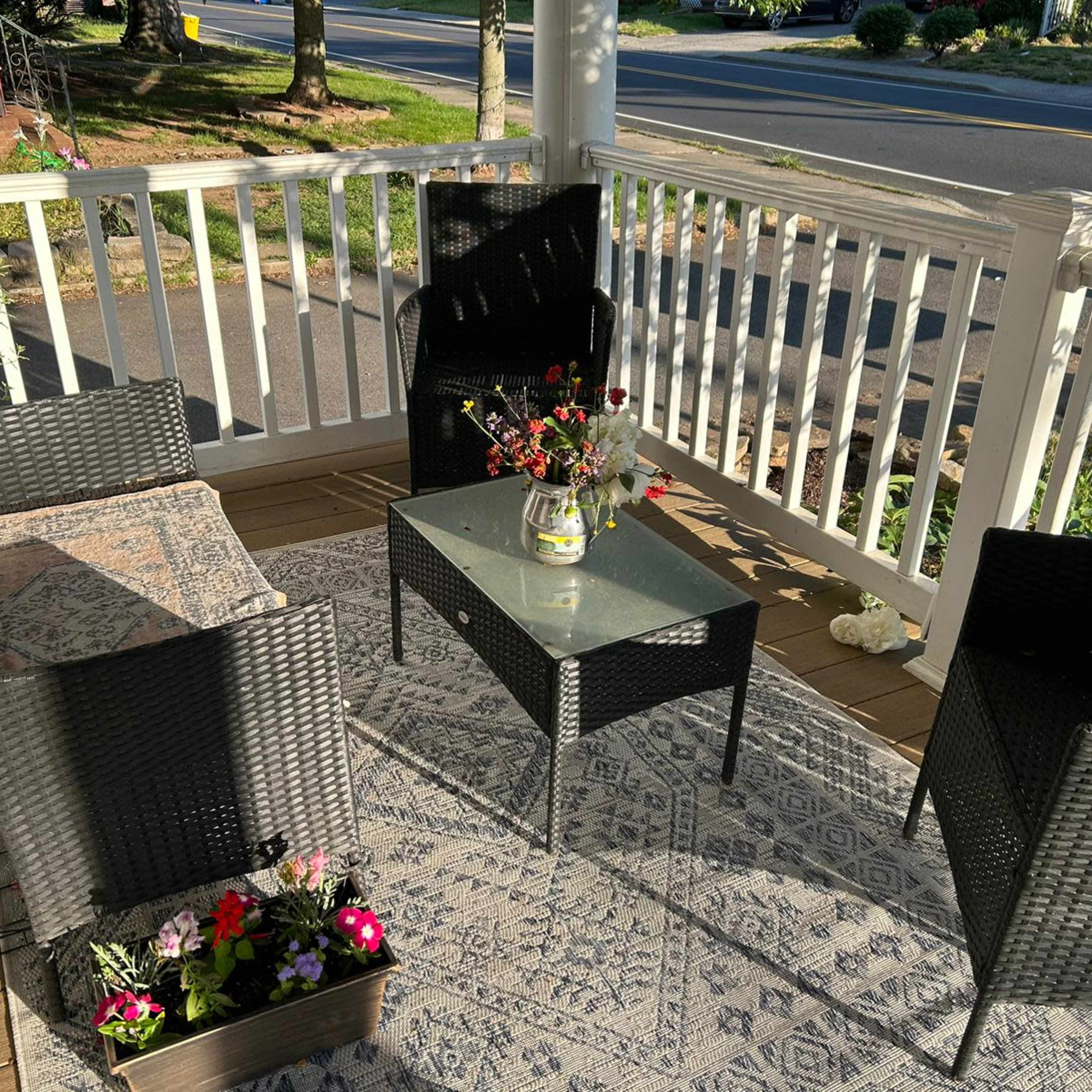 A porch features four black wicker chairs around a glass table, with flowers in a vase and planters on a patterned rug.