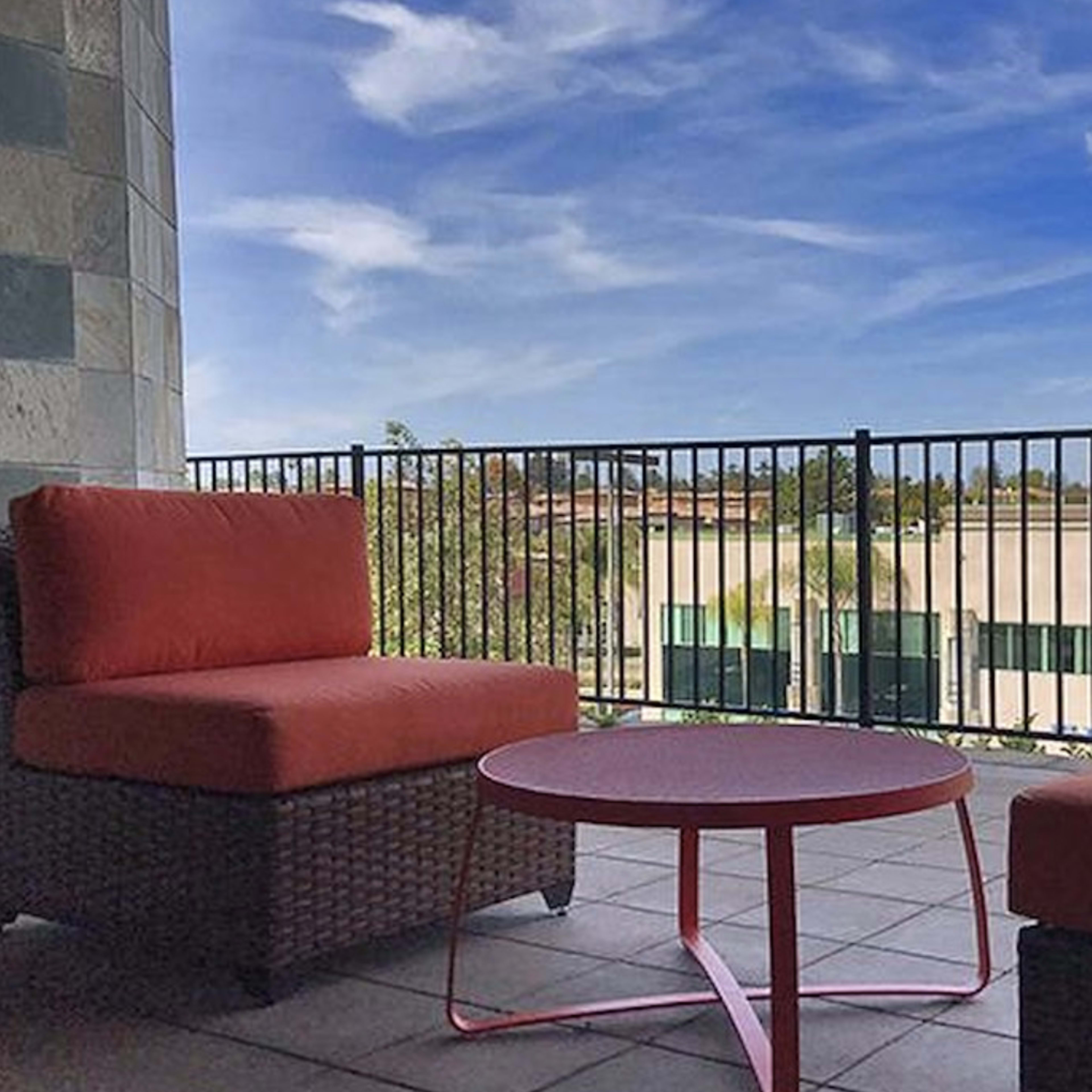 A balcony featuring two red cushioned chairs and a round table, overlooking a clear blue sky and a building in the background.