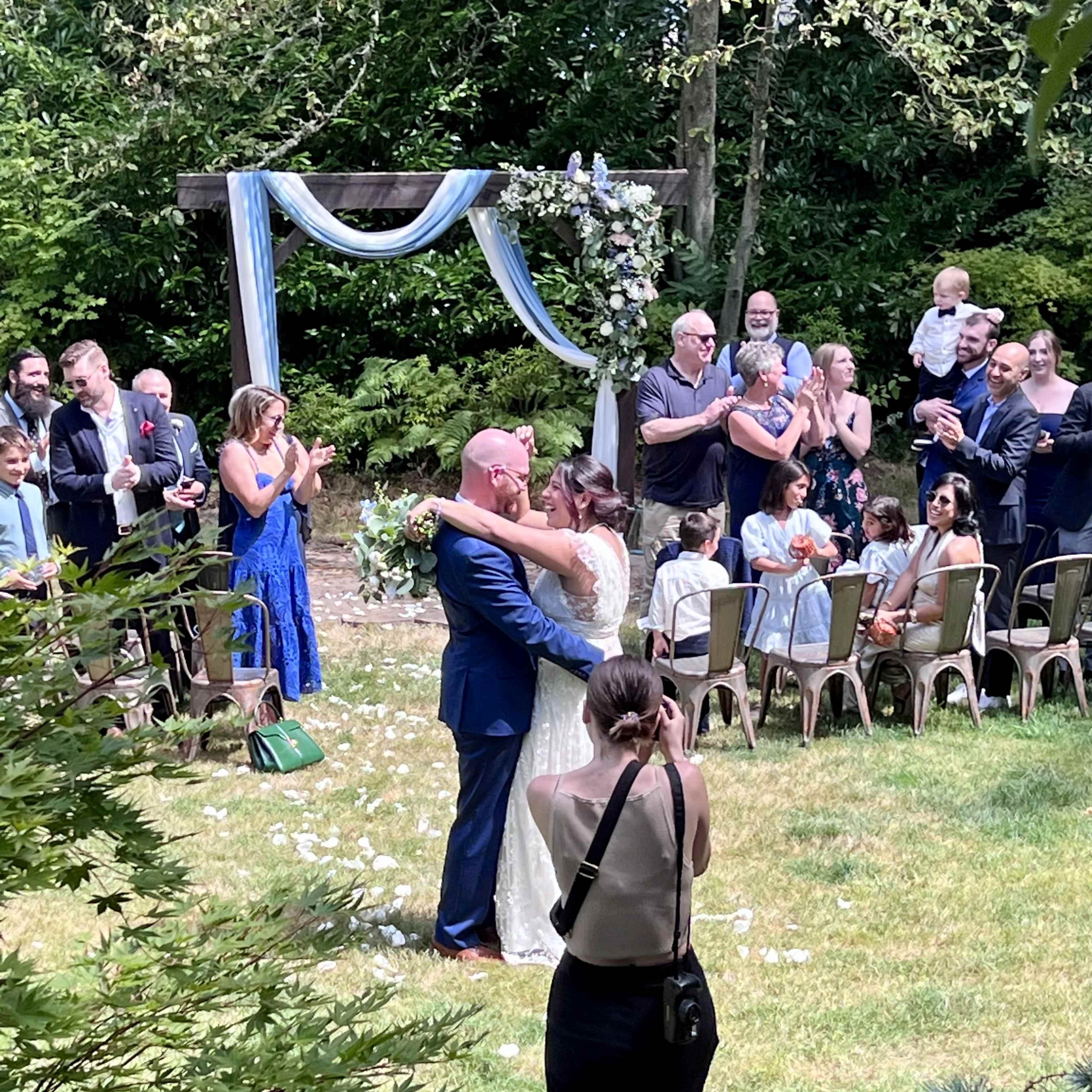 A couple exchanges a kiss during their outdoor wedding ceremony while guests applaud nearby.