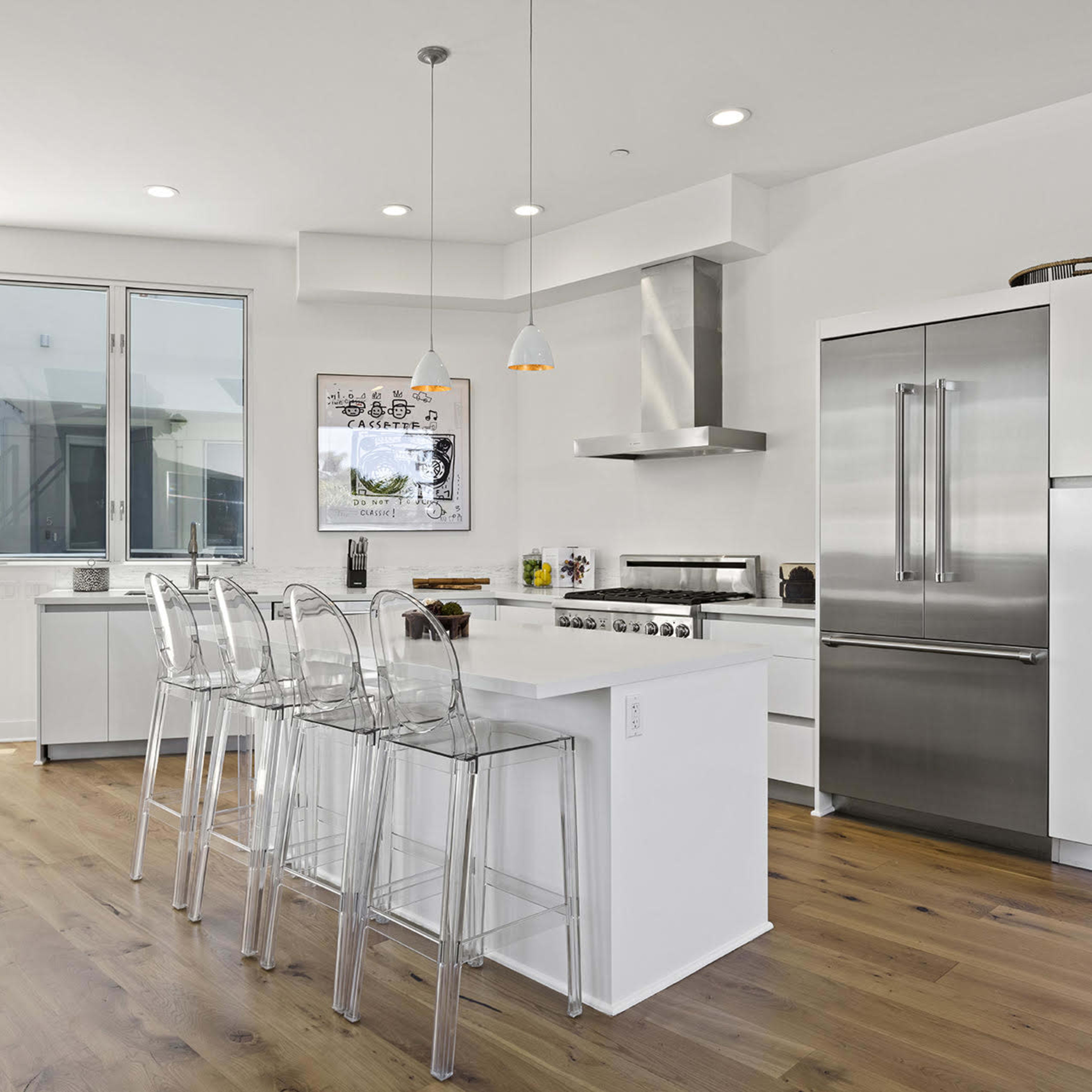 A modern kitchen features stainless steel appliances, a large island with clear bar stools, and floor-to-ceiling windows that open to a balcony.