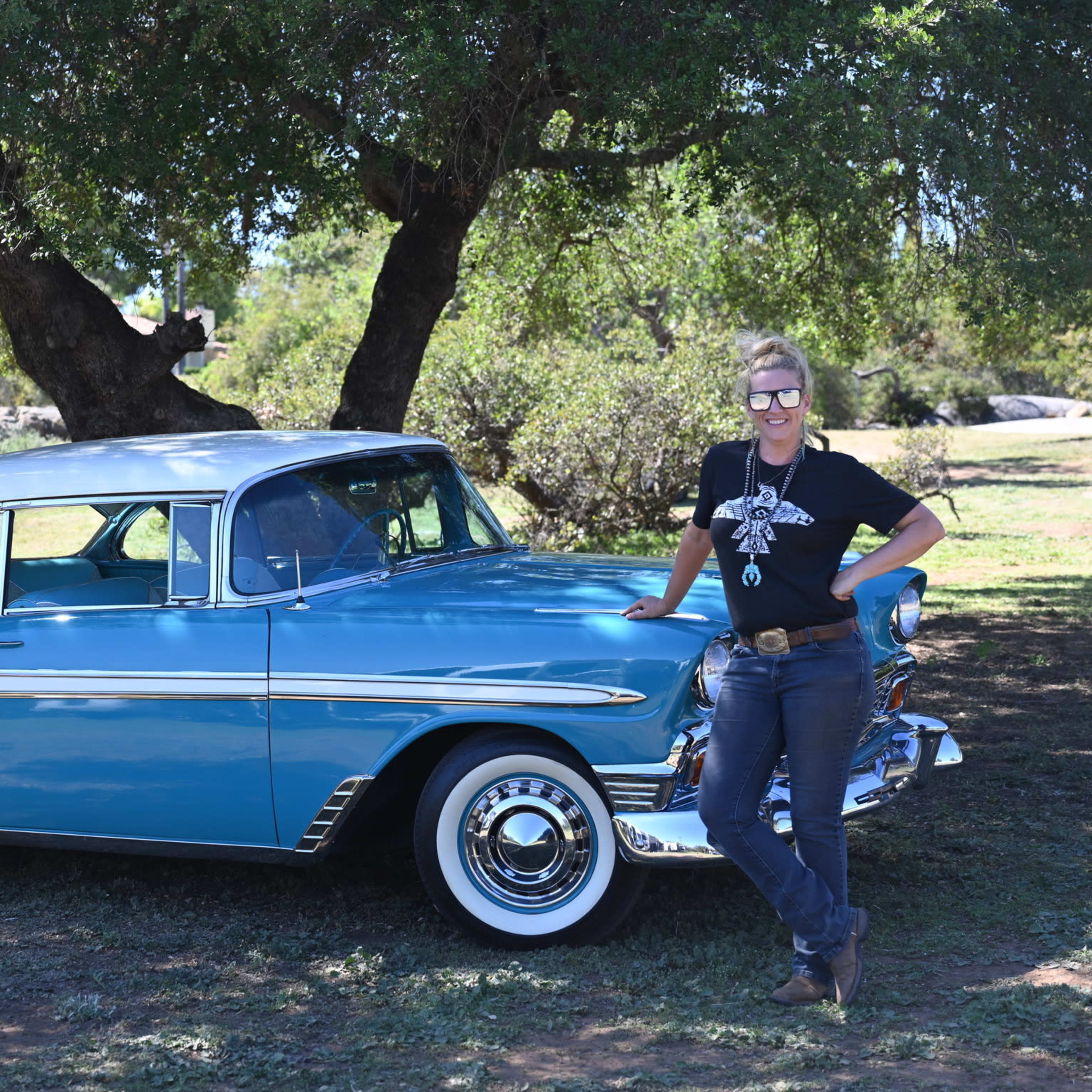 A woman stands beside a vintage blue and white car parked under a large tree in a grassy area.