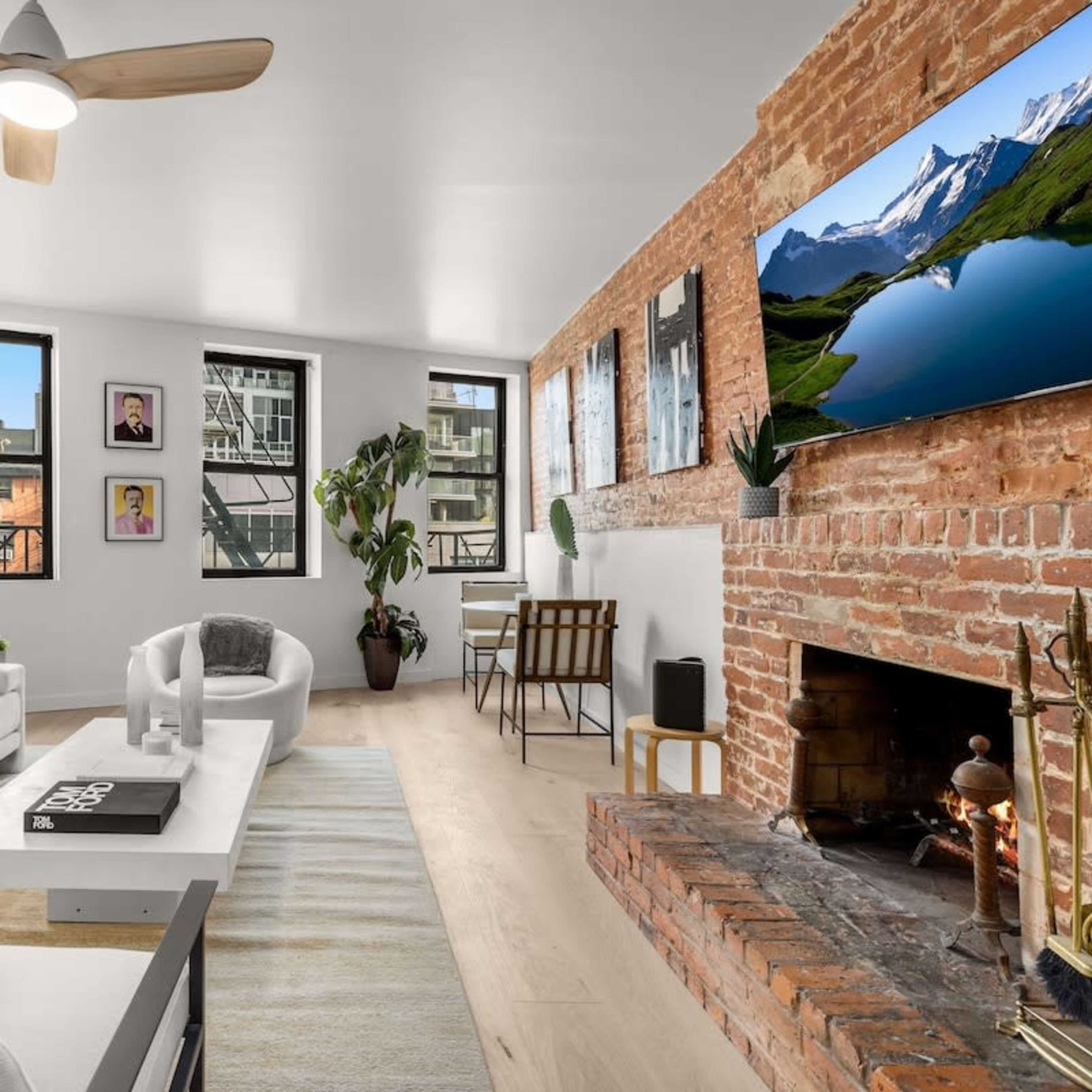 A modern living room with a white sectional sofa, a coffee table, a brick fireplace, and large windows allowing natural light, adorned with framed artwork and a flat-screen TV mounted on the wall.