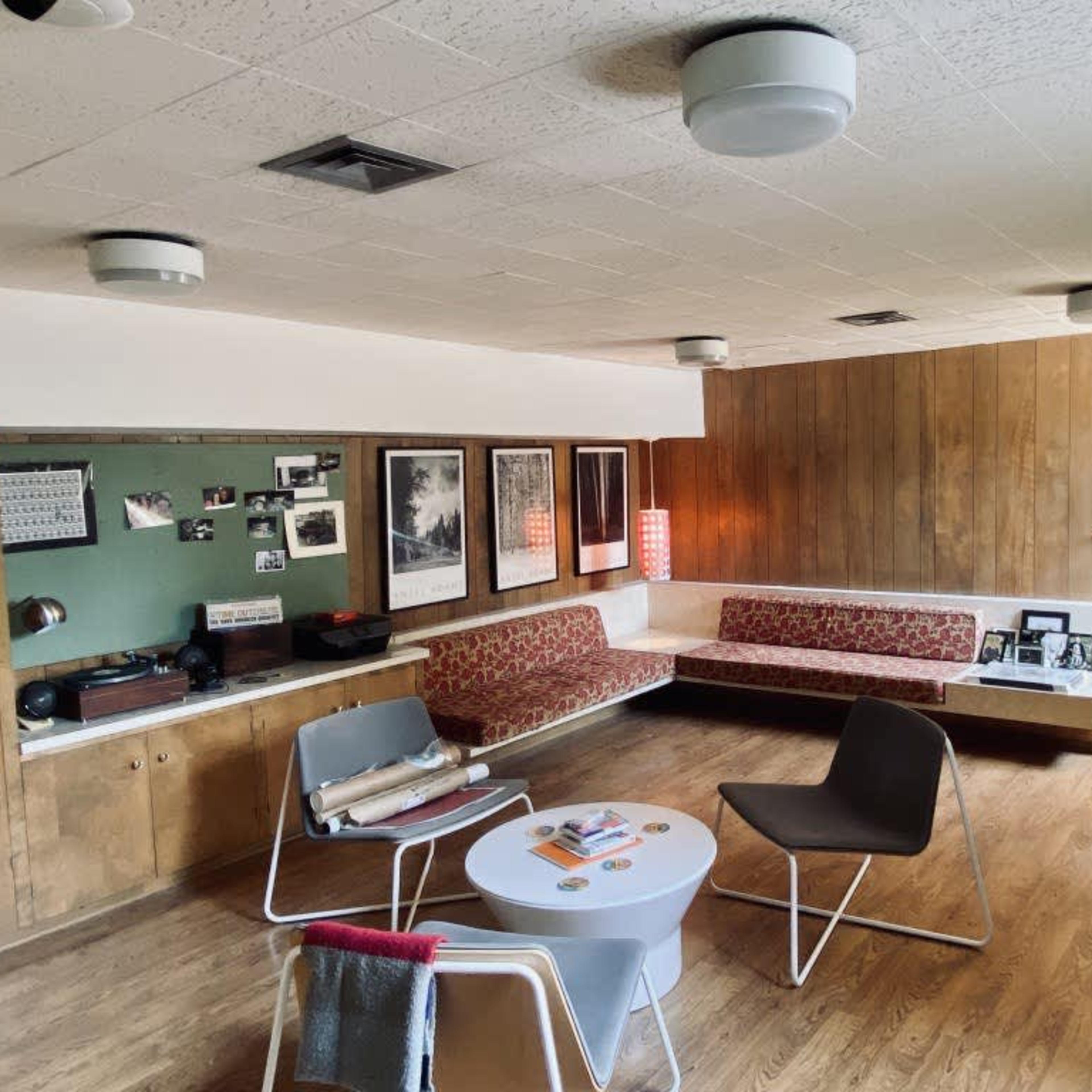 A mid-century modern living room with wooden paneling, a sectional sofa, a round coffee table, and various framed artworks and photographs on the walls.