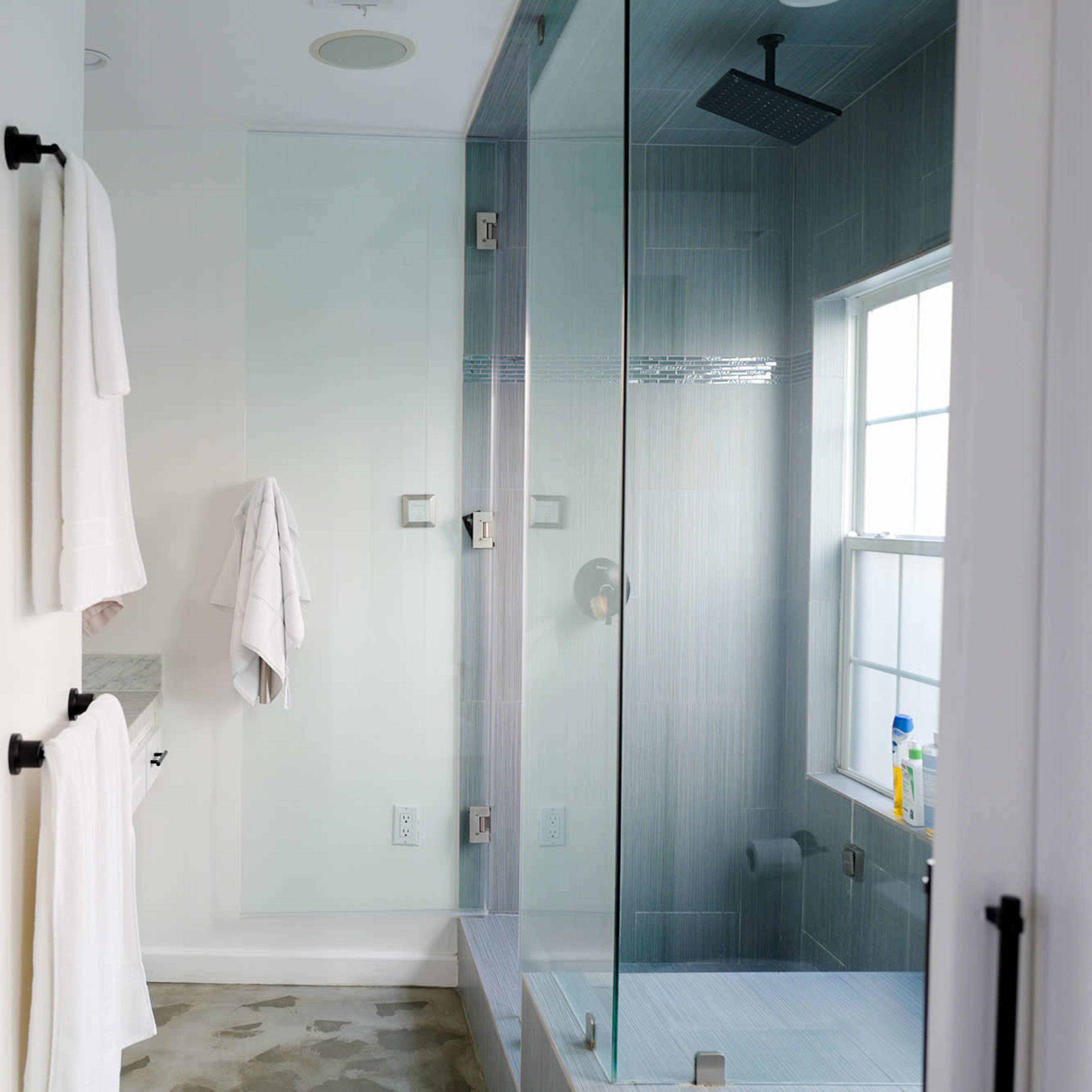 A modern bathroom features a glass shower enclosure, white towels hanging on racks, and a window allowing natural light.