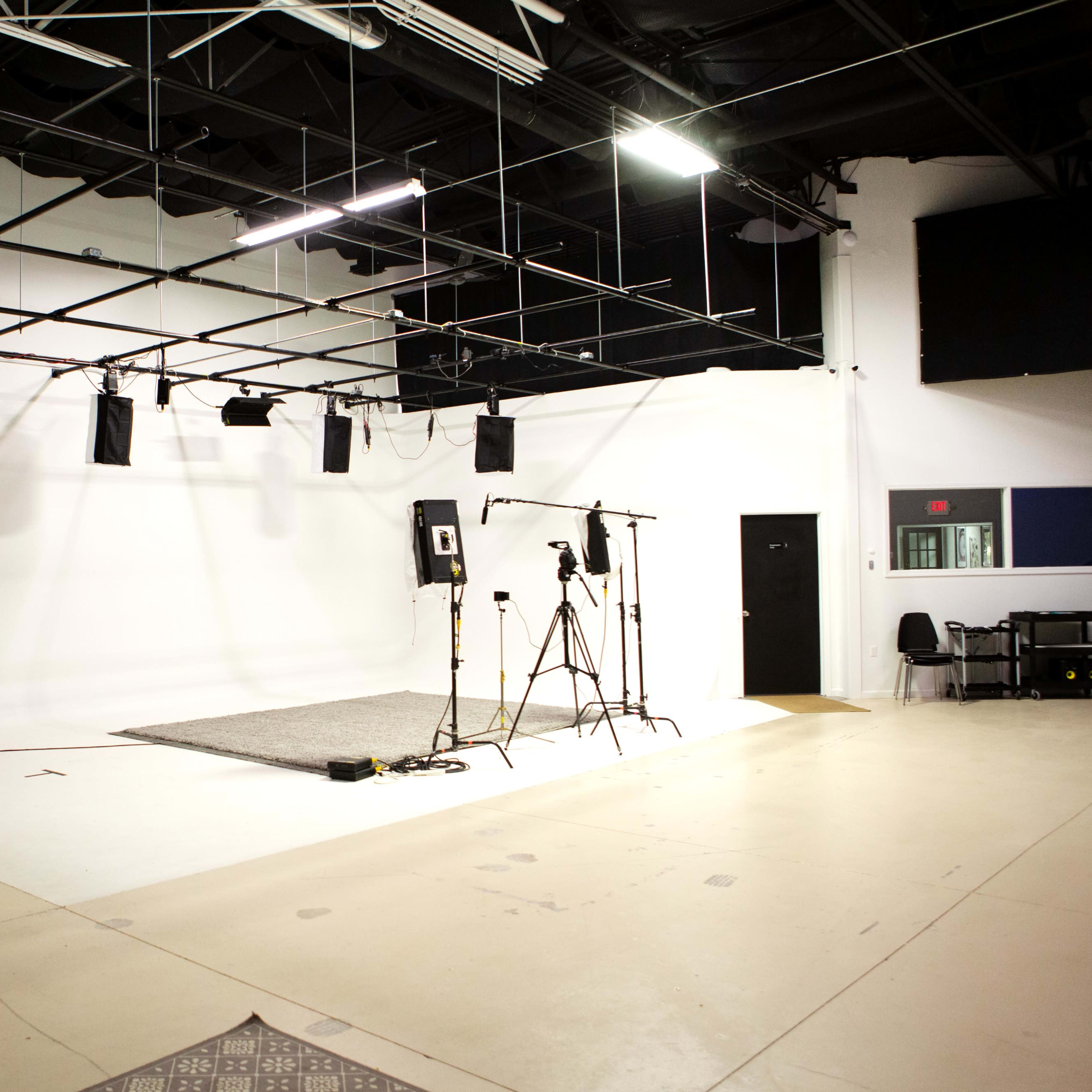 A sparse studio space featuring a gray rug, several lights on tracks, and camera equipment set up against a blank wall.