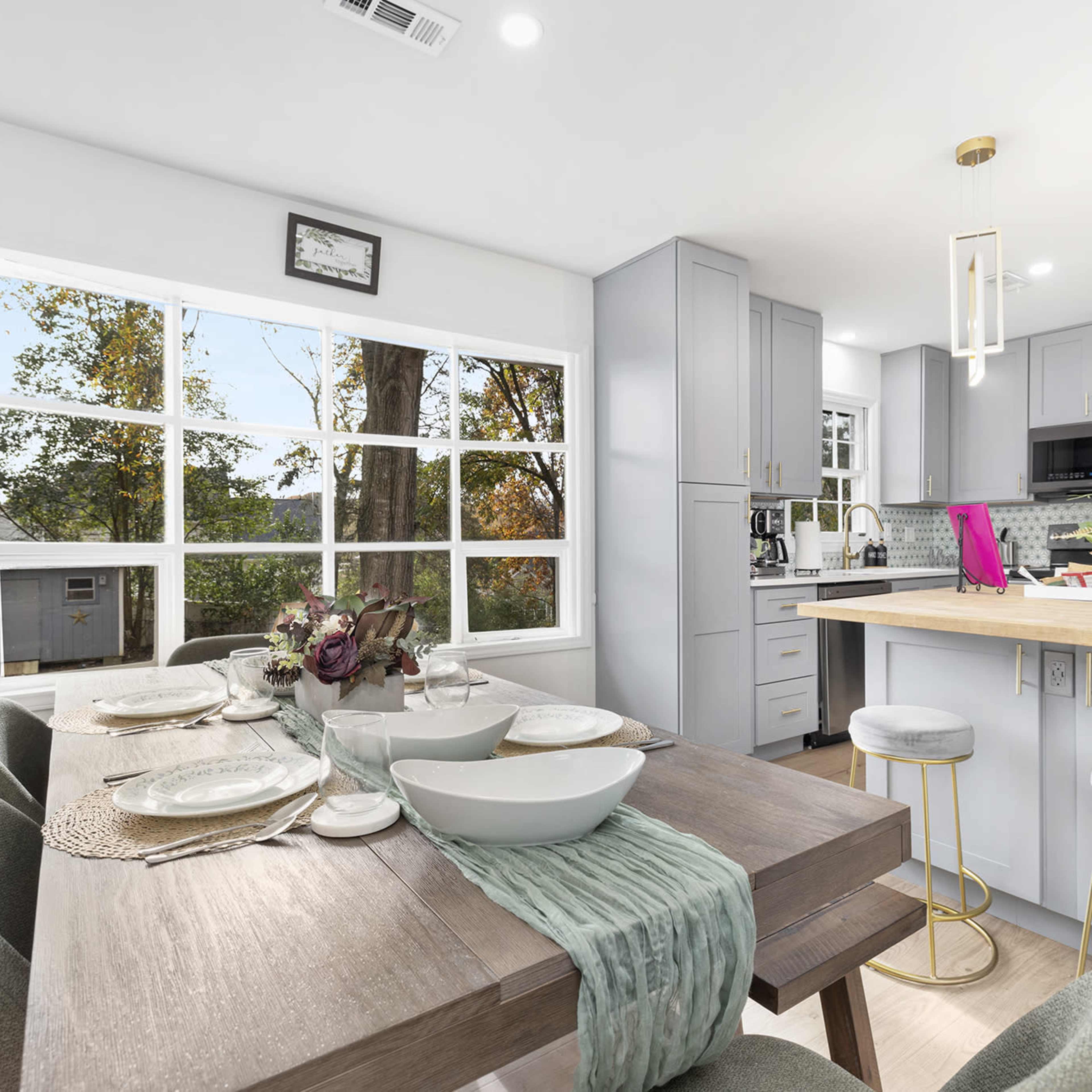 A modern kitchen and dining area feature a wooden table set for dinner, with a light-colored island and stainless steel appliances in the background.