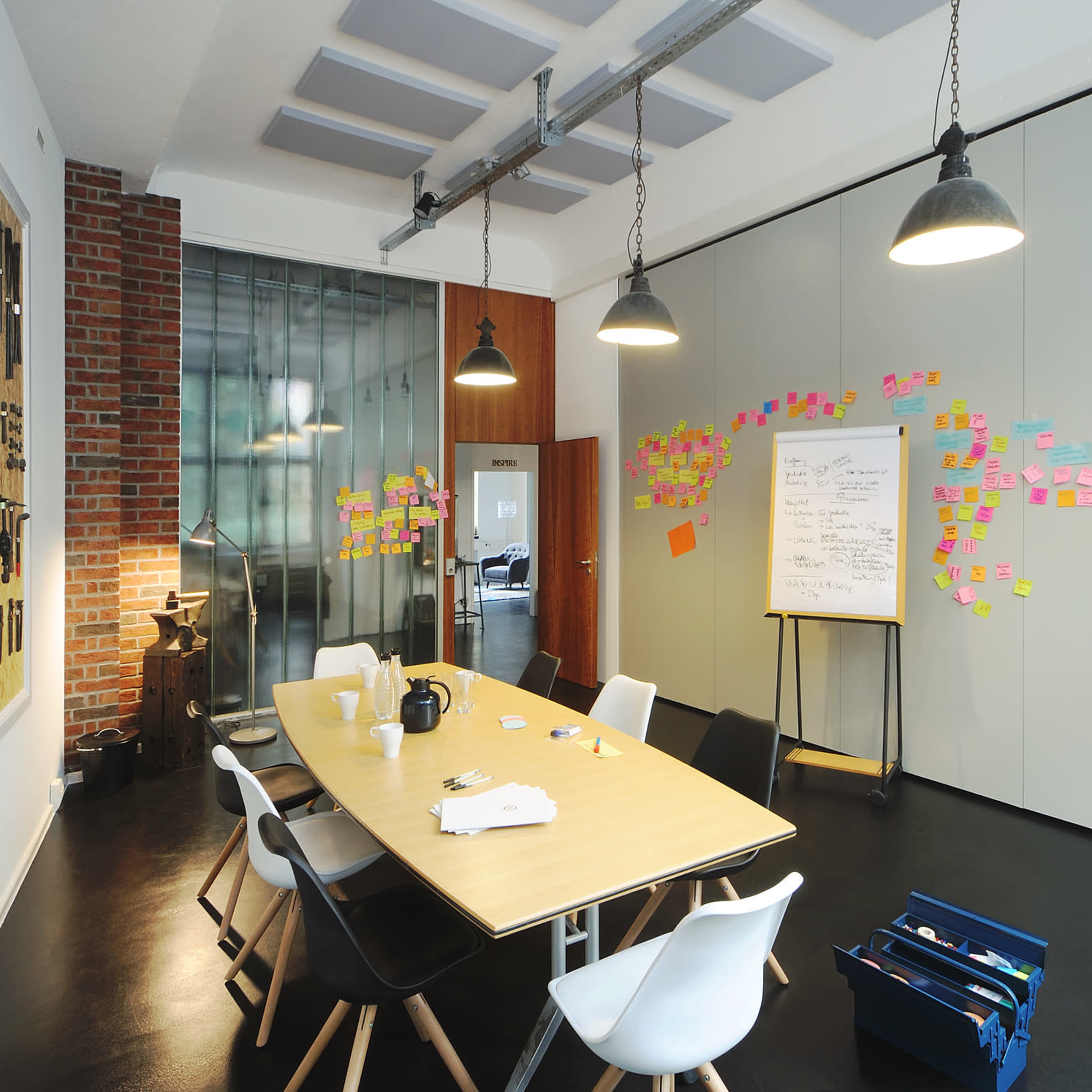 A modern meeting room features a large table surrounded by chairs, with a wall of tools and colorful sticky notes arranged on the adjacent walls.