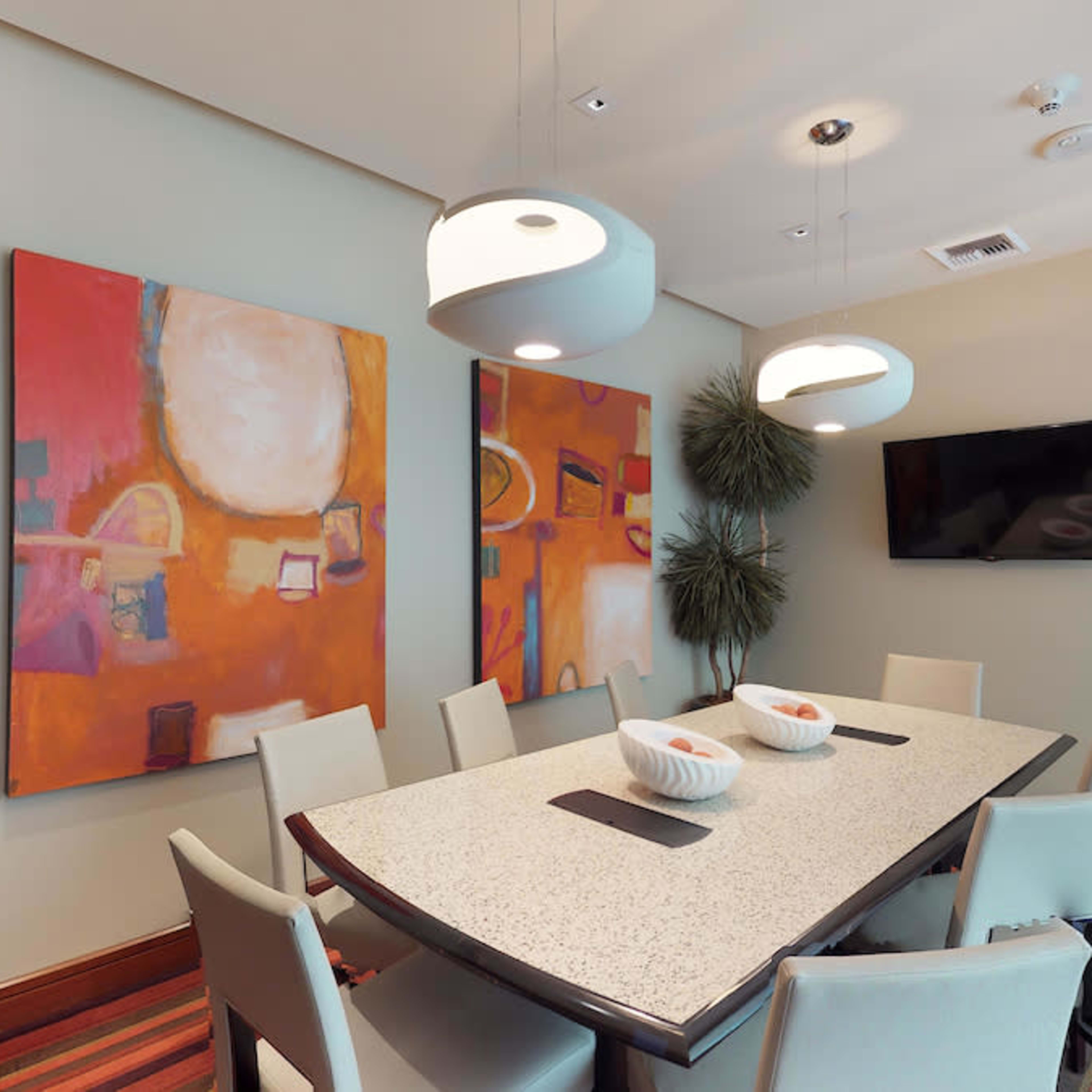 A dining room features a large table surrounded by chairs, with two abstract paintings on the walls and pendant lights overhead.
