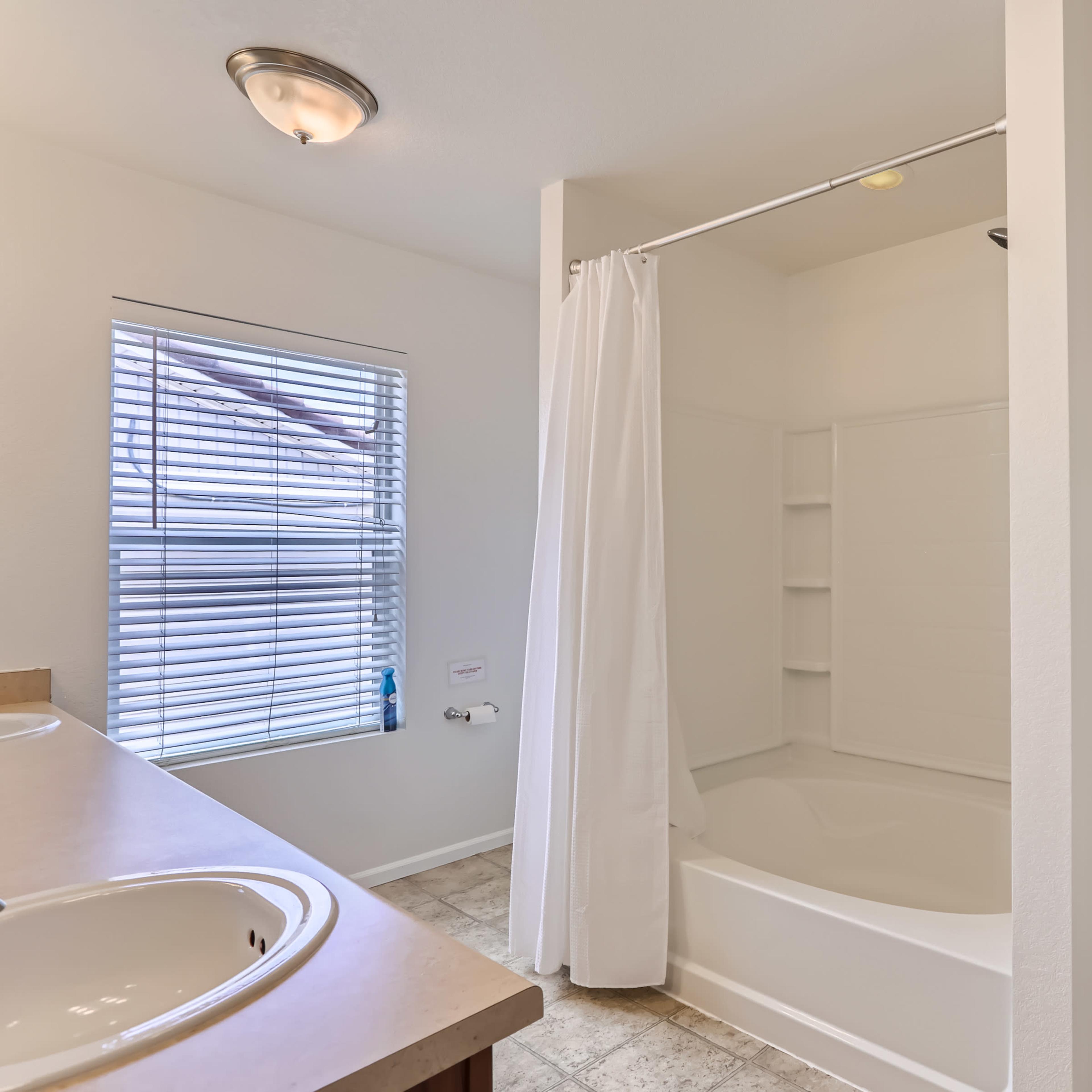 A clean bathroom featuring a shower with a curtain, a double sink vanity, a window with blinds, and shelving stocked with folded towels.