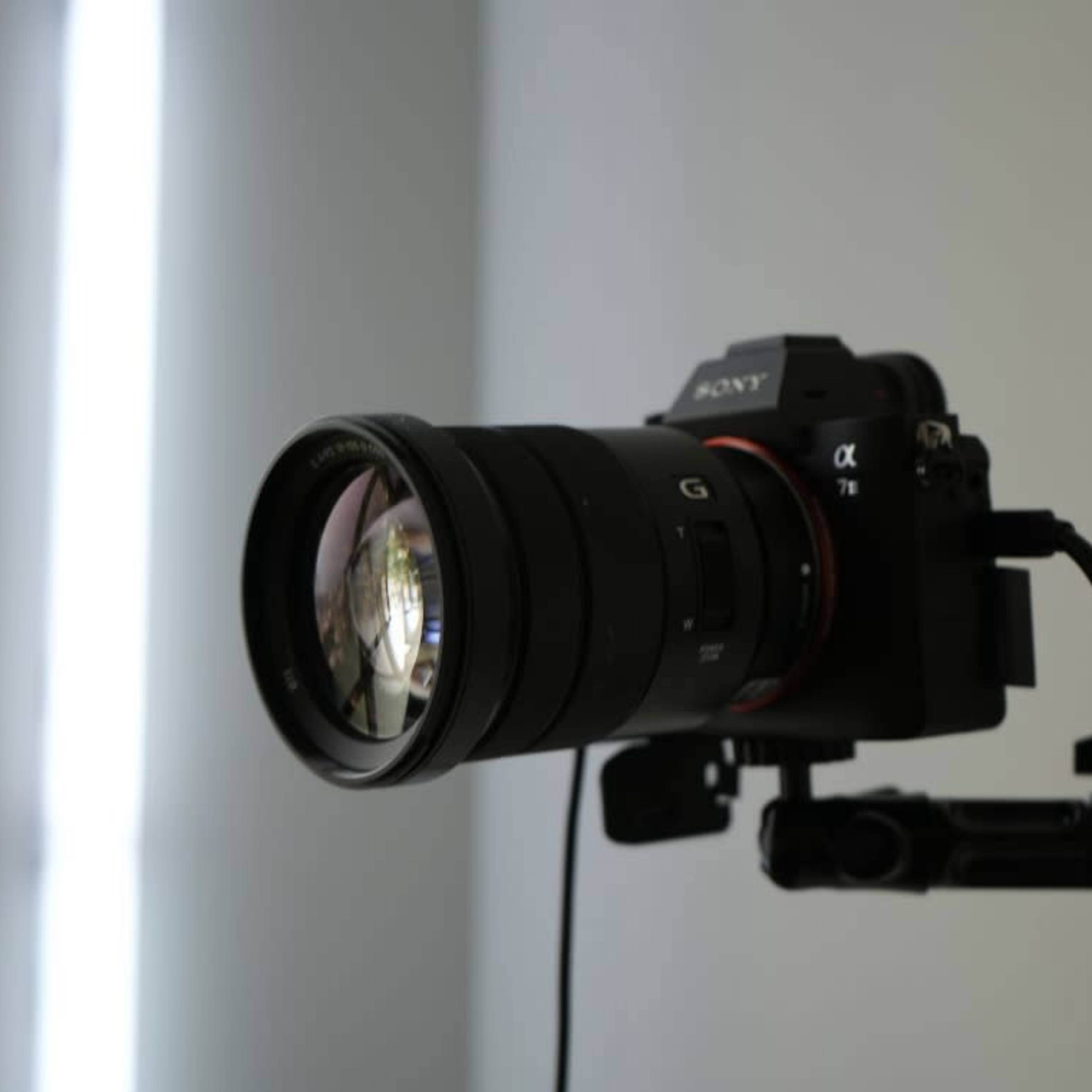 A Sony Alpha camera with a large lens is mounted on a stable support unit against a plain background.