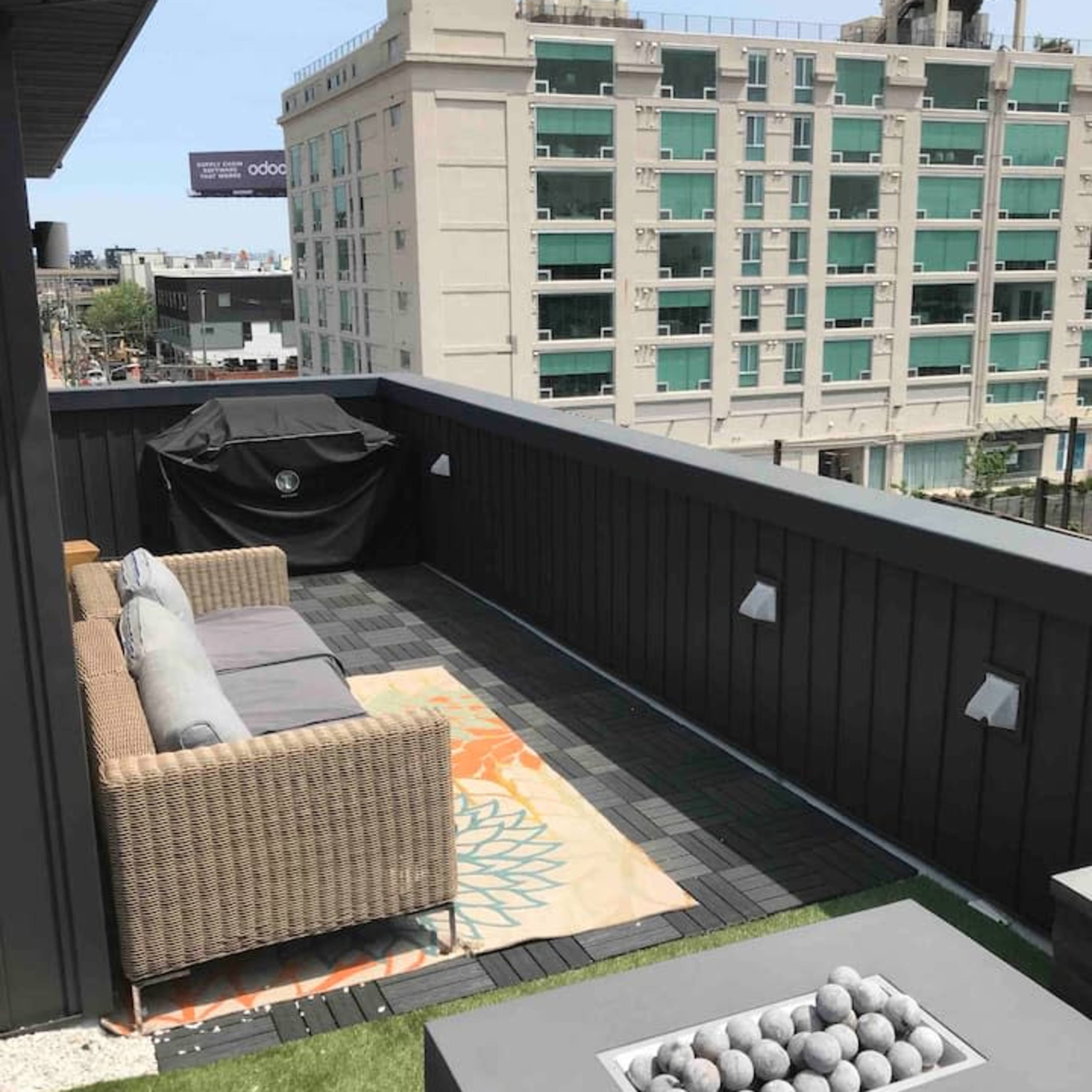 A rooftop terrace with a sofa, decorative rug, and a fire pit surrounded by green grass and city buildings in the background.