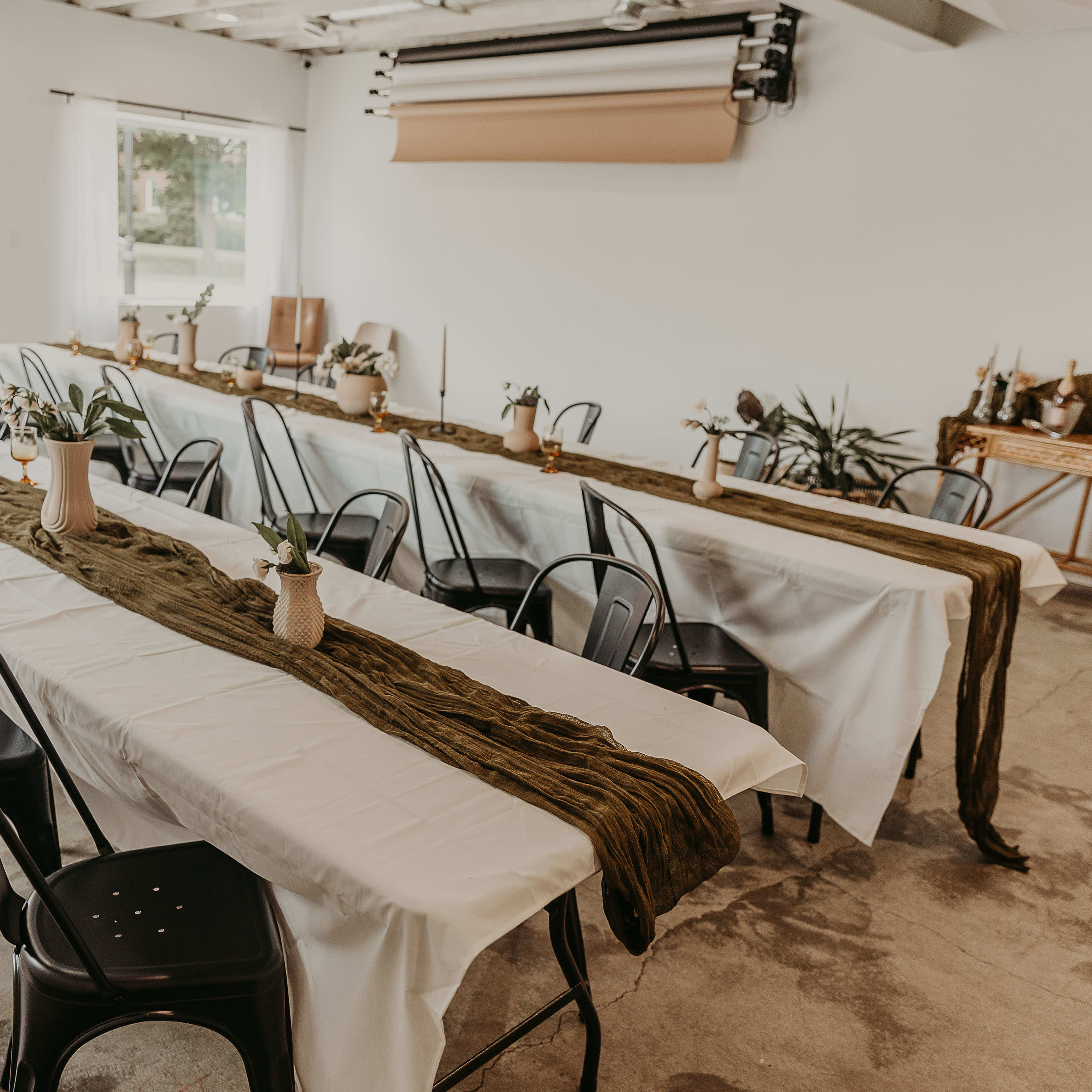A spacious room features several long tables set with white tablecloths, centerpieces of potted plants, and decorative items along a green runner.